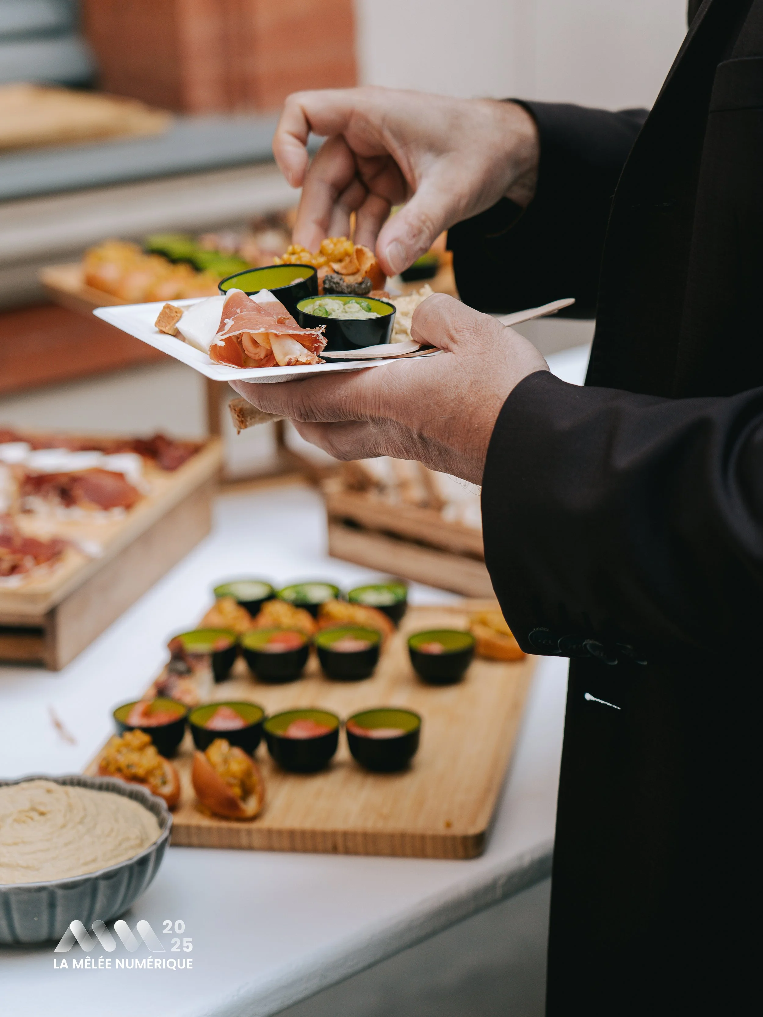 Personne tenant une assiette de tapas avec diverses bouchées, devant une sélection de nourriture sur un buffet, y compris des petits bols de sushi et une assiette de charcuterie.