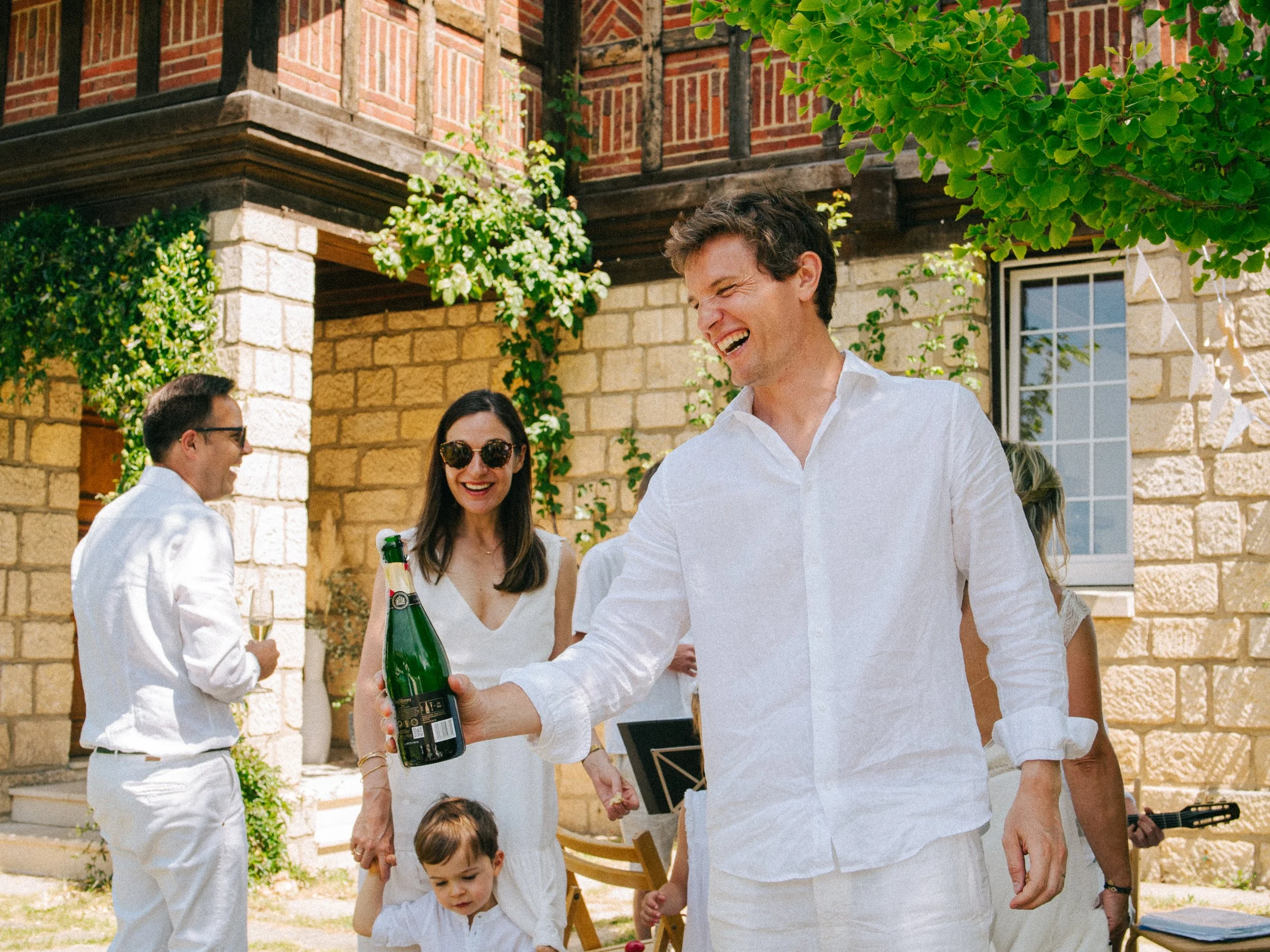 Un groupe de personnes habillées en blanc lors d'une fête en plein air, tenant des boissons et souriant, avec un bâtiment en pierre en arrière-plan.
