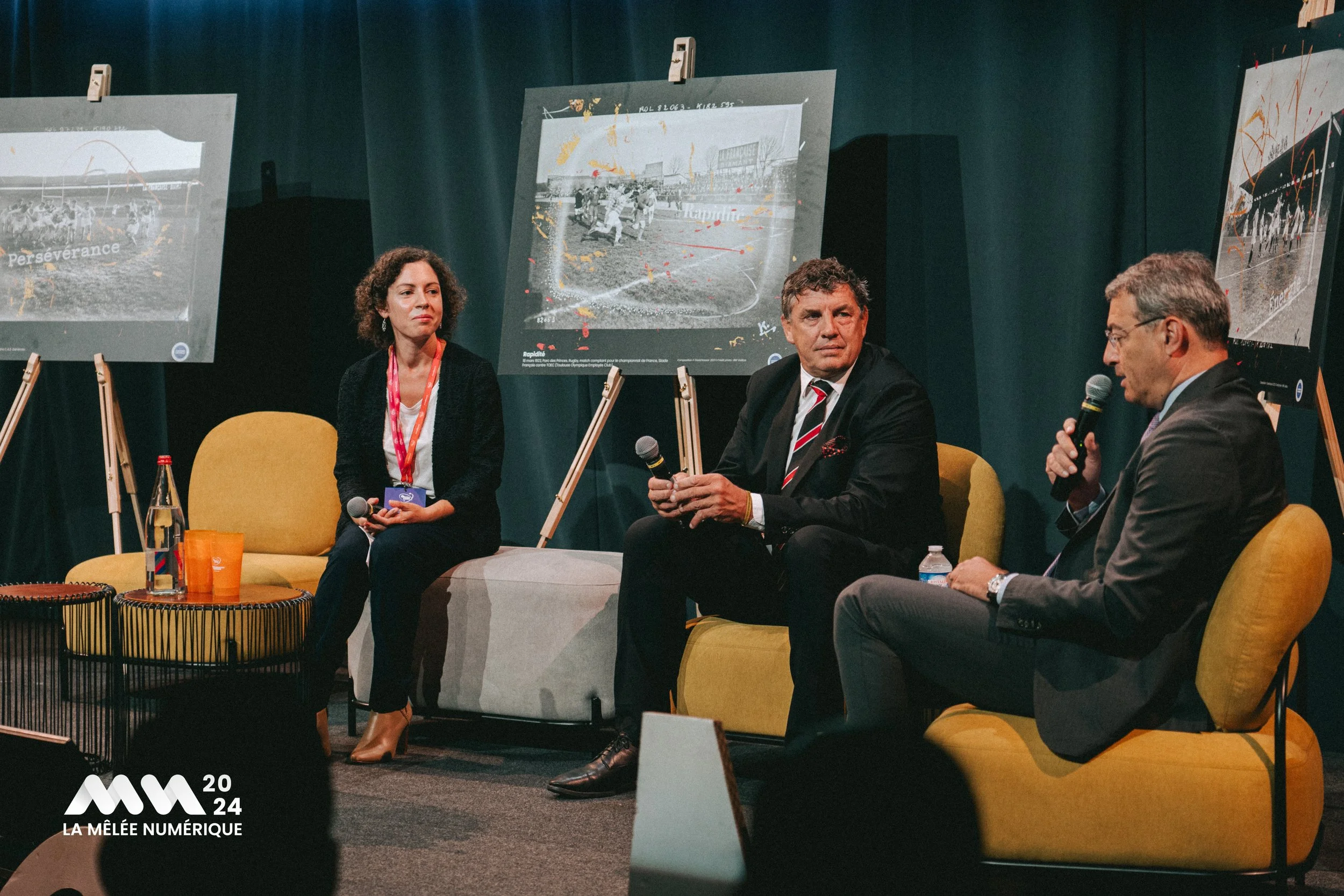Trois personnes assises lors d'une conférence, avec des photographies sur des chevalets derrière elles. Il y a une femme et deux hommes, tous avec un micro à la main. La scène semble se dérouler en 2024 à La Mêlée Numérique.
