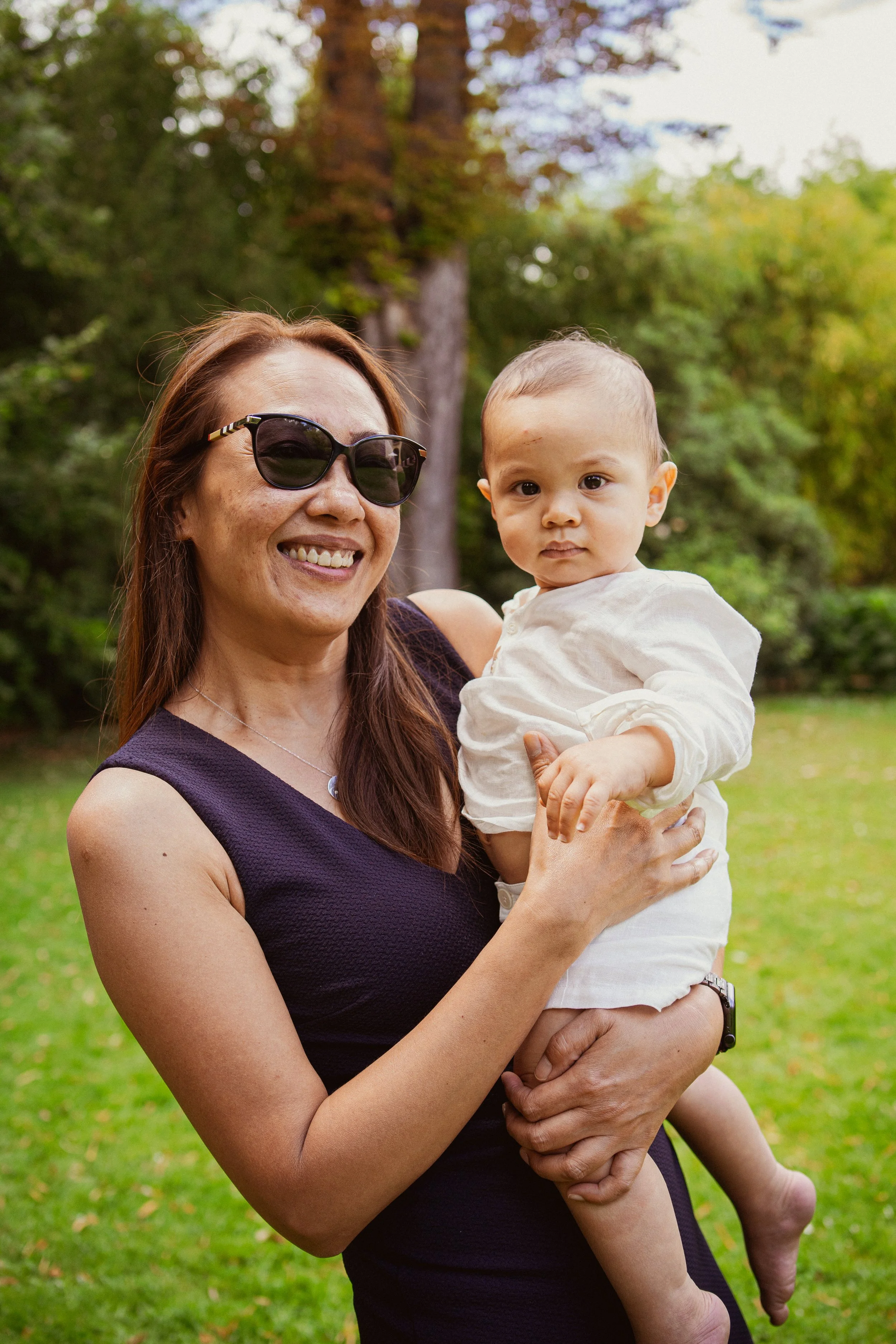 Une femme souriante portant des lunettes de soleil, tenant un bébé dans ses bras, dans un parc avec des arbres en arrière-plan.