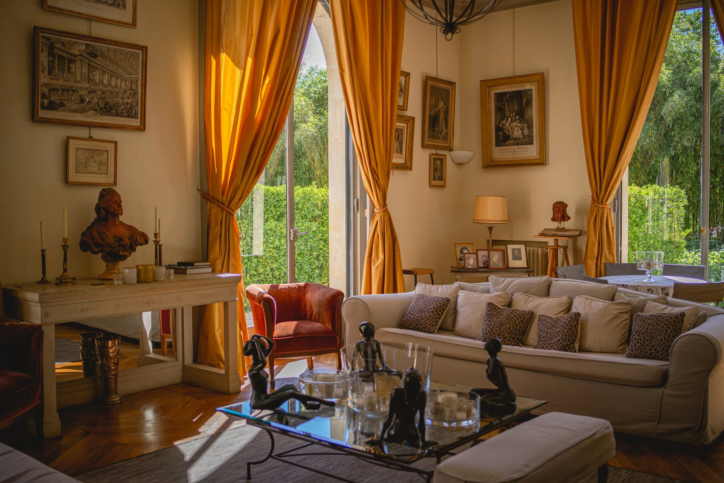 Salon élégant avec canapé blanc, coussins, tableau, sculptures, et grandes fenêtres avec rideaux dorés laissant entrer la lumière naturelle, décorés de photos de famille et d'œuvres d'art.