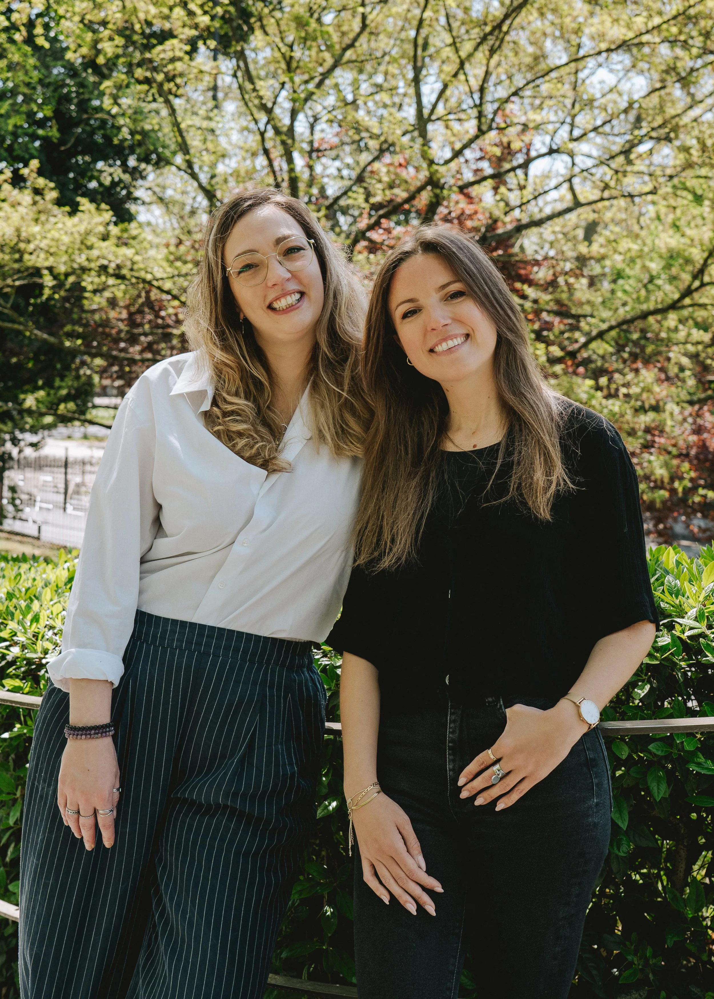 Deux femmes souriantes posent dehors devant des arbres verts en pleine nature, l'une porte une chemise blanche et un pantalon rayé, l'autre une veste noire et un jean noir.