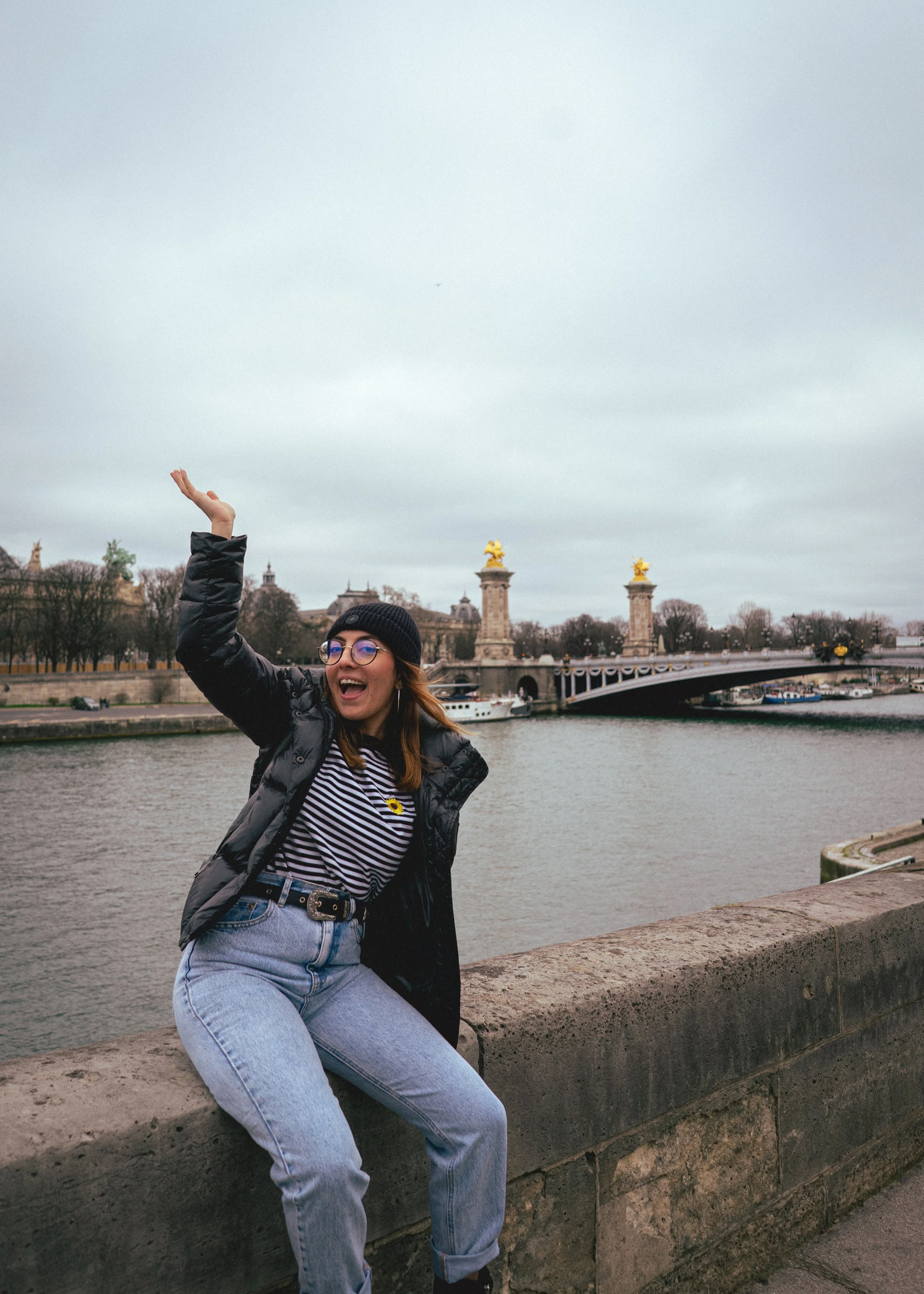 Une femme souriante avec un bonnet noir, portant un blazer en jean, un t-shirt rayé, et des lunettes, assise sur un mur en pierre le long d'une rivière, avec un pont avec des statues dorées en arrière-plan.