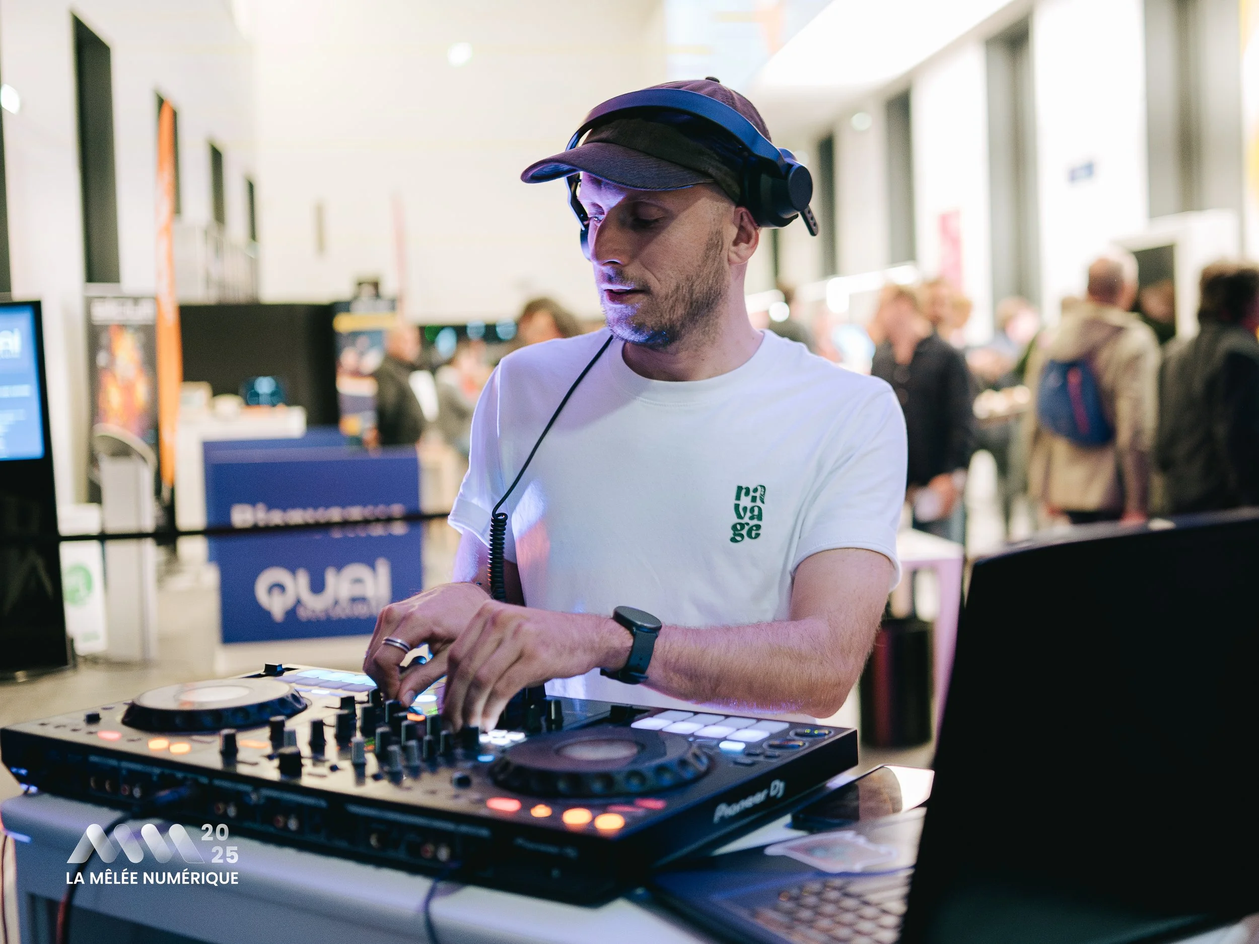 Un DJ homme portant un t-shirt blanc, un casque et une montre, utilisant un contrôleur DJ dans une salle en intérieur lors d'un événement en 2025.
