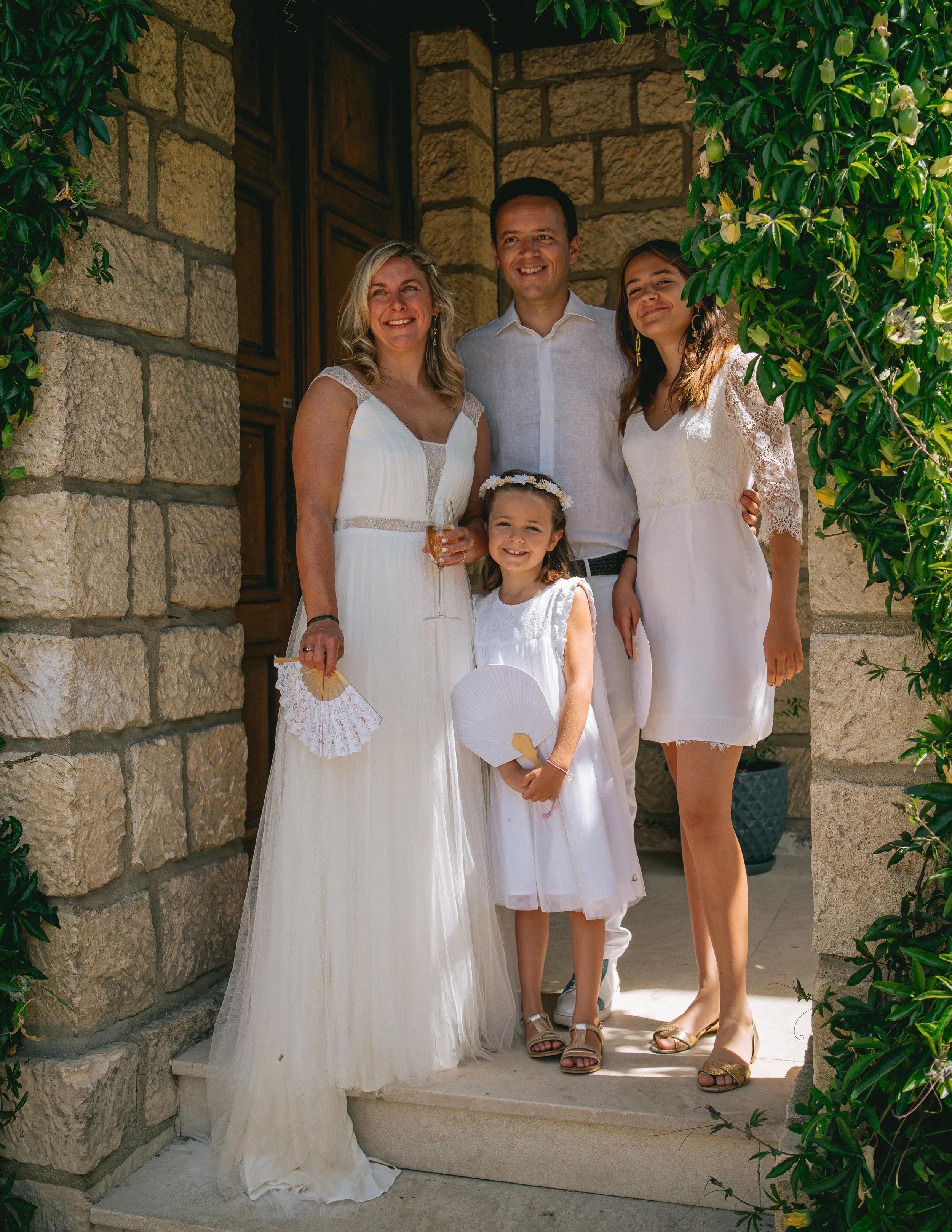 Groupe de cinq personnes debout devant une porte en pierre, portant des vêtements blancs lors d'une occasion spéciale, avec deux jeunes filles et trois adultes, entourés de végétation luxuriante.
