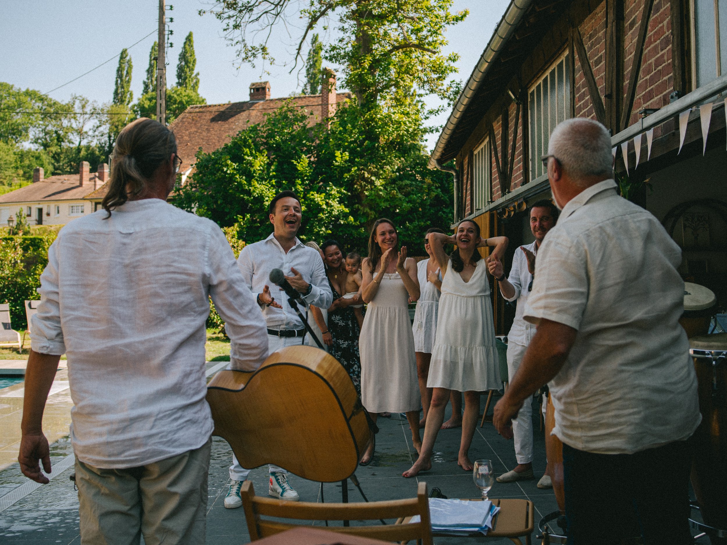 Un groupe de personnes souriantes dans une fête en plein air, avec des musiciens jouant de la guitare, dans un jardin lors d'une journée ensoleillée.