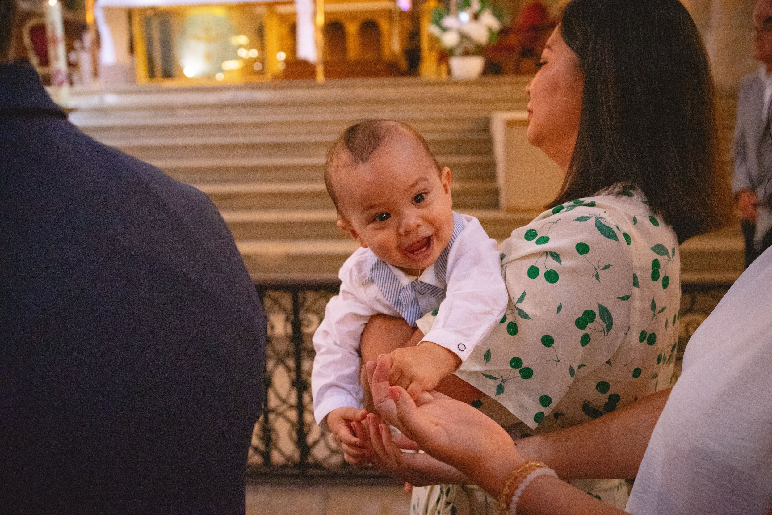 Une femme tient un bébé souriant dans une église, avec des personnes en arrière-plan.