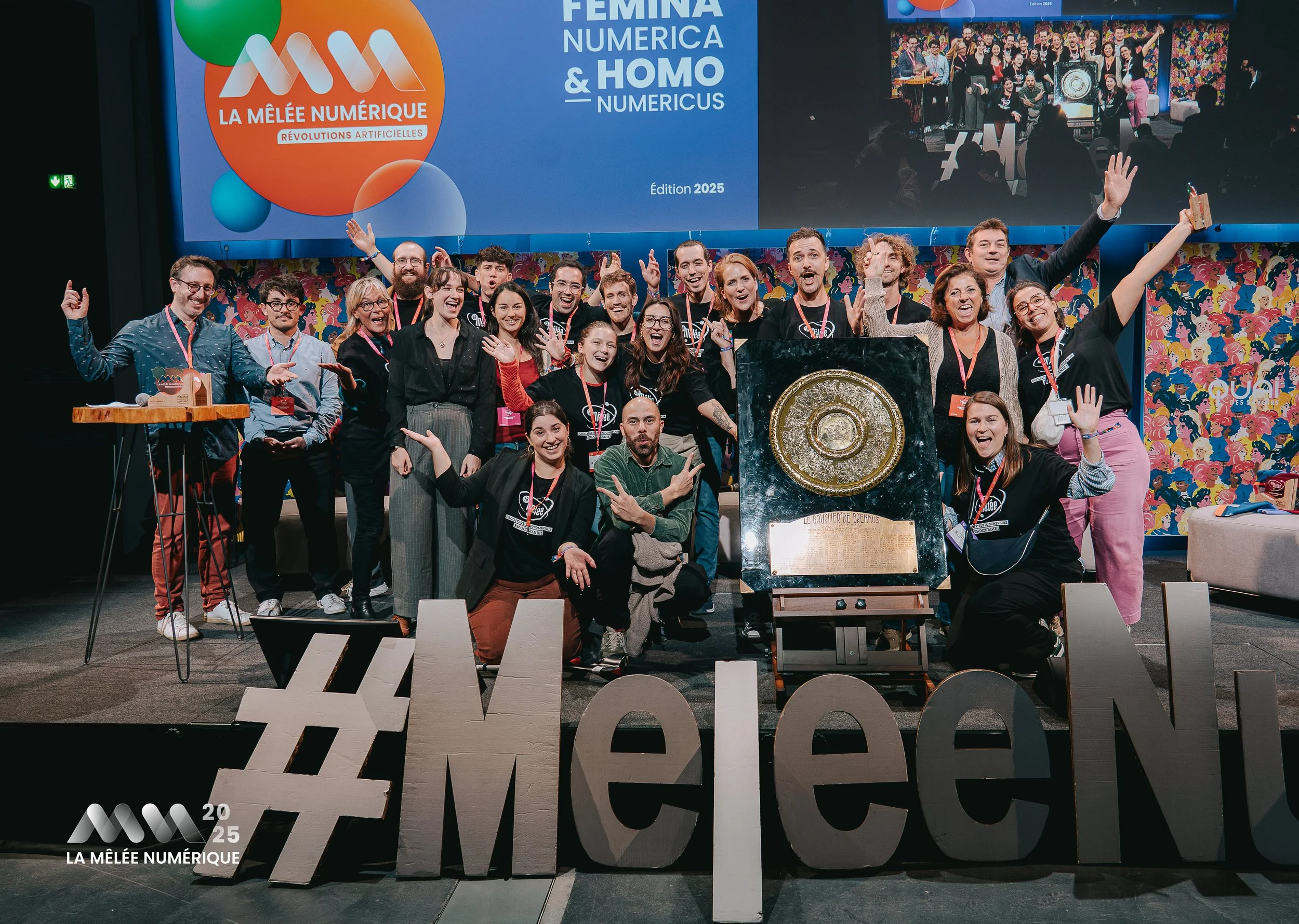 Groupe de personnes posant avec un trophée lors d'un événement appelé La Mêlée Numérique 2025, avec un grand hashtag #MêleeN en avant, à l'arrière-plan un écran avec le texte 'La Mêlée Numérique' et 'Femina, Numérica & Homo Numéricus, Édition 2025'.