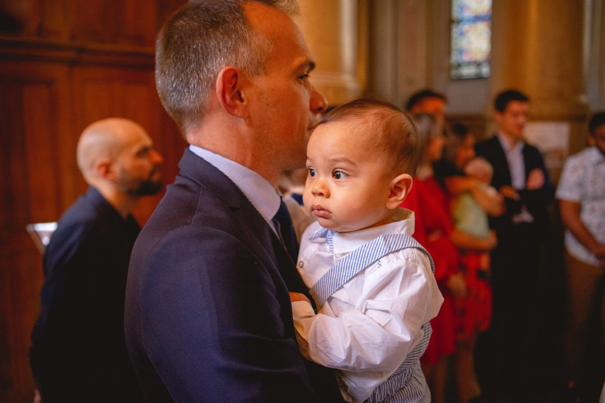 Un homme en costume blanc tient un jeune enfant dans ses bras lors d'un événement religieux ou cérémonie dans une église.