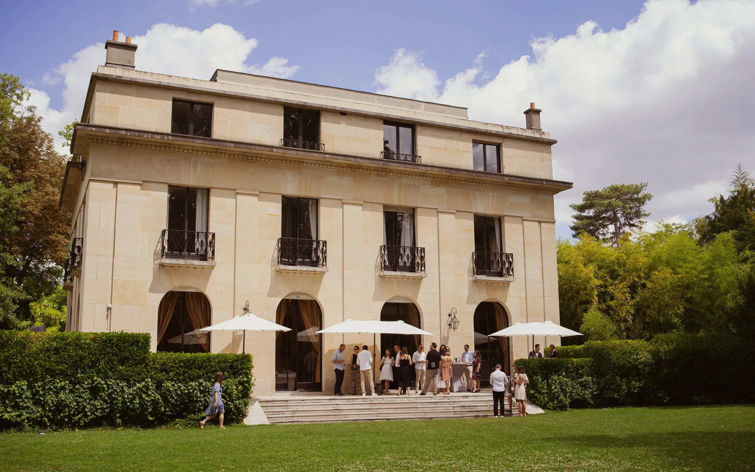Une grande maison en pierre beige avec des fenêtres à petits balcons en fer forgé noir. Un groupe de personnes habillées élégamment est rassemblé sur la terrasse, sous des parasols blancs, lors d'un événement en plein air. Il y a un espace vert bien 