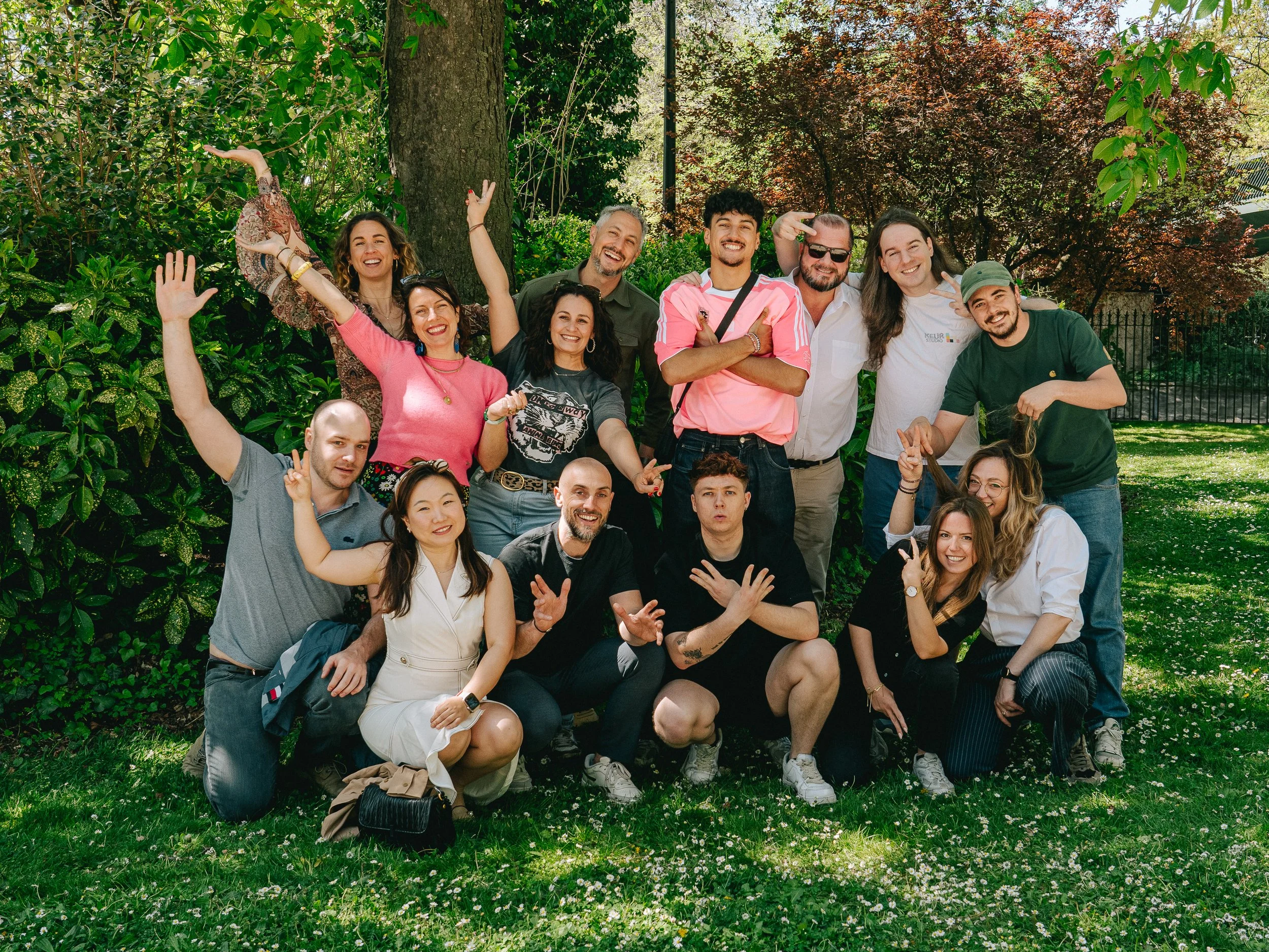Groupe de personnes souriantes et souriantes en plein air, debout et assises, entourées d'arbres et d'arbustes verts, posant pour une photo de groupe en journée ensoleillée.