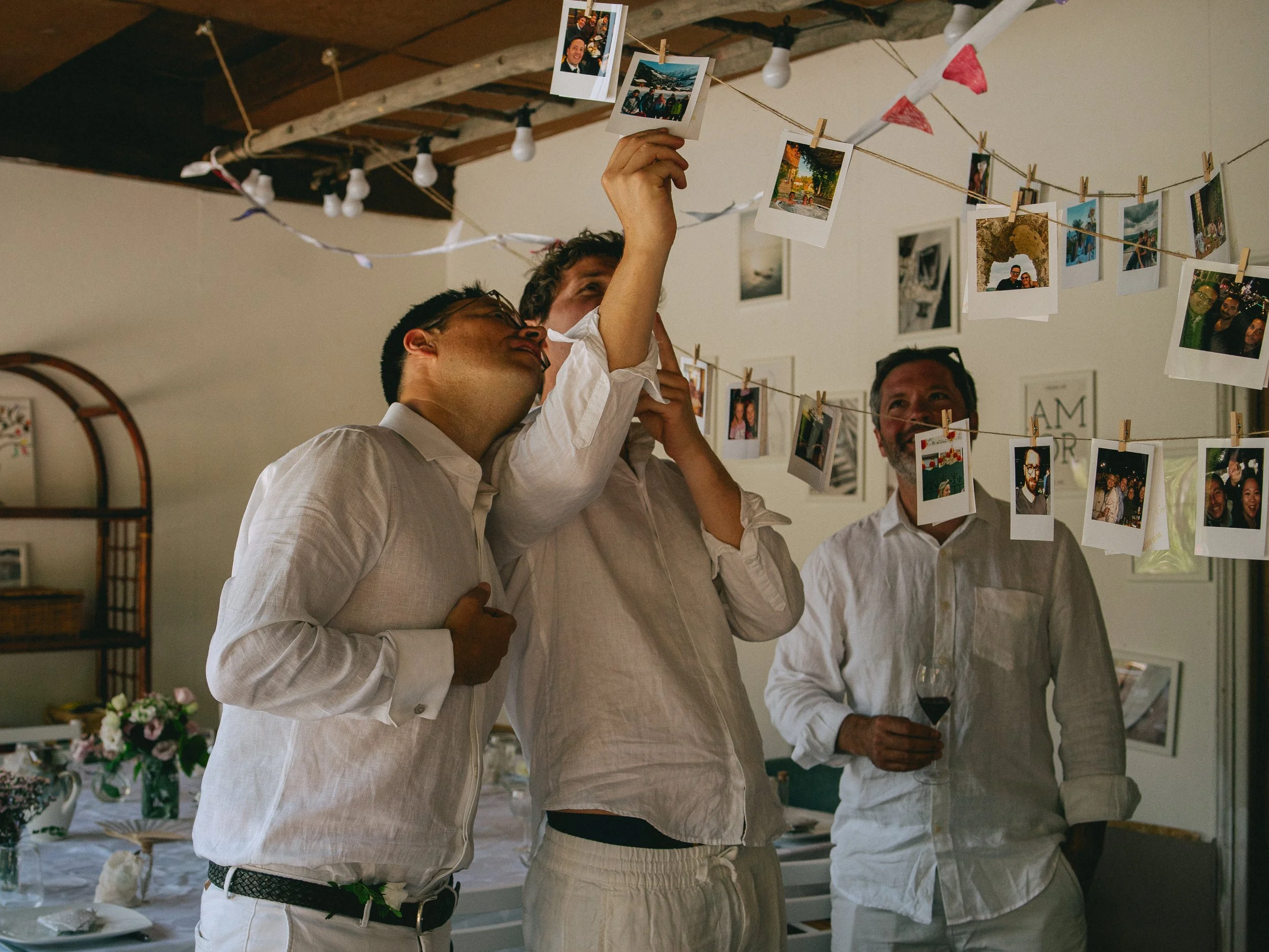 Trois hommes en chemise blanche décorent une pièce avec des photos accrochées sur une corde à linge, lors d'une fête ou d'un rassemblement.