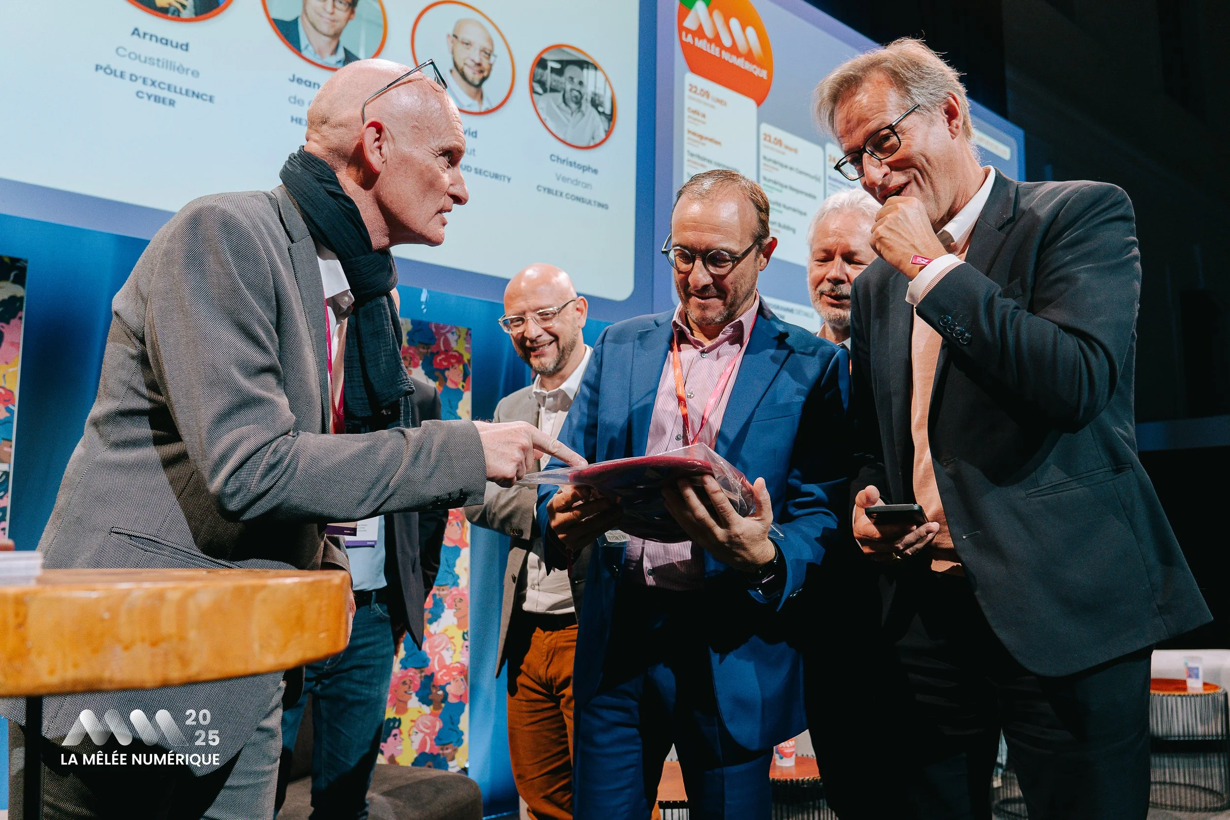 Groupe de cinq hommes en costume discutant à une conférence, avec un grand écran en arrière-plan montrant une présentation.