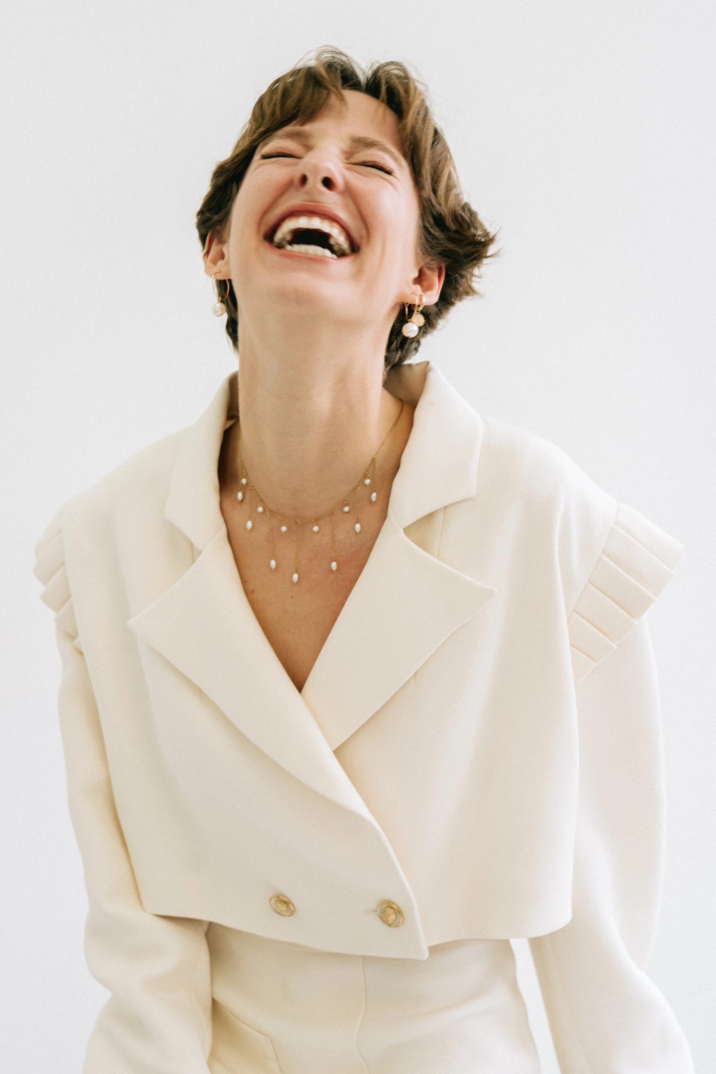 Femme souriante portant une veste blanche, avec des boucles d'oreilles et un collier en perles, tête légèrement inclinée en arrière.
