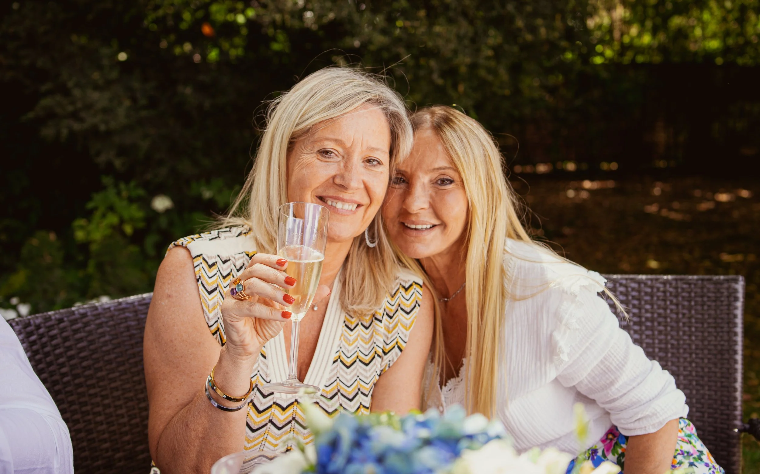 Deux femmes souriantes à une fête en extérieur, l'une tenant une flûte de champagne.