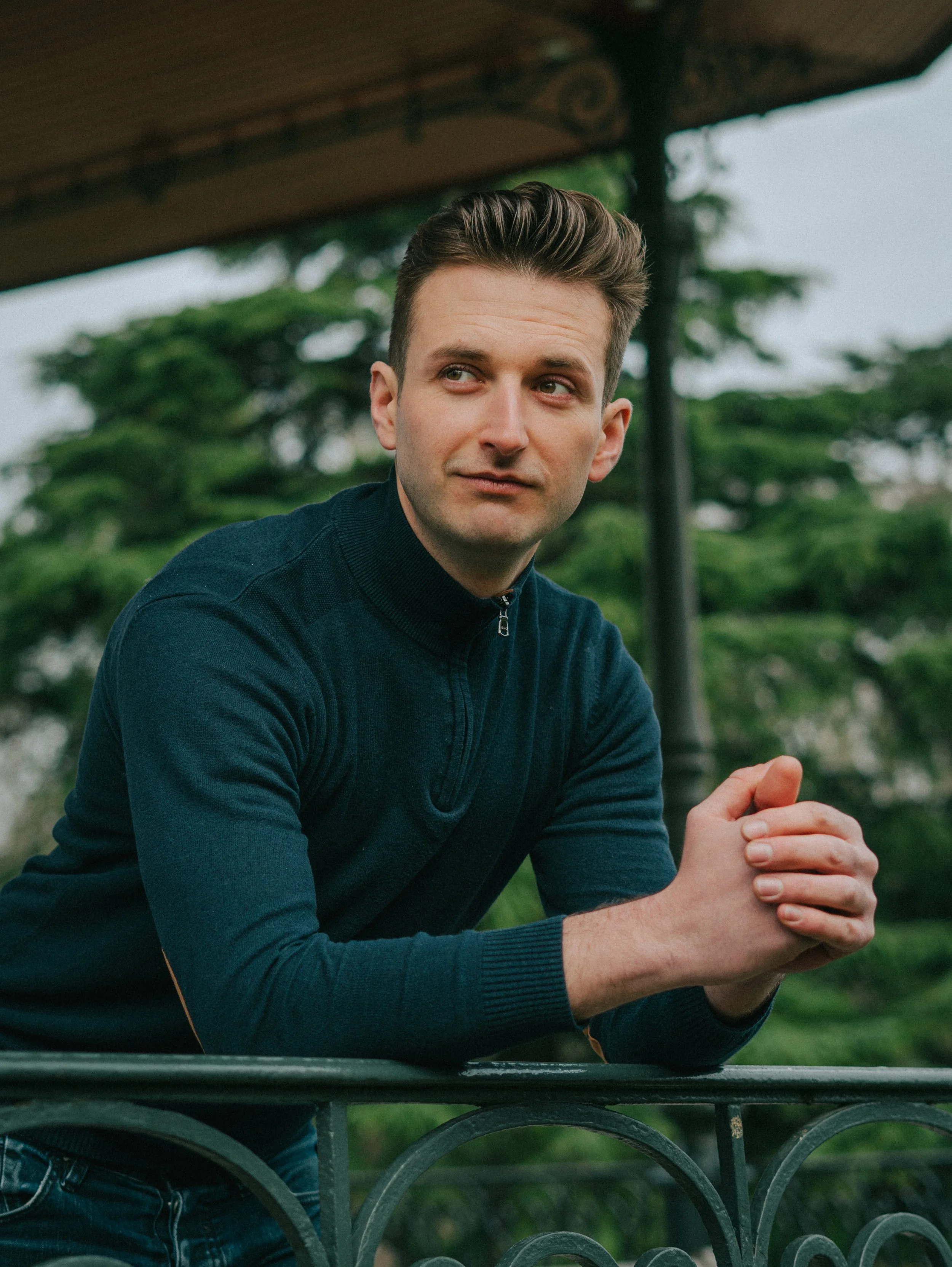 Jeune homme avec cheveux bruns, portant un pull en col montant noir, appuyé sur une barrière en fer, dans un parc avec des arbres en arrière-plan.