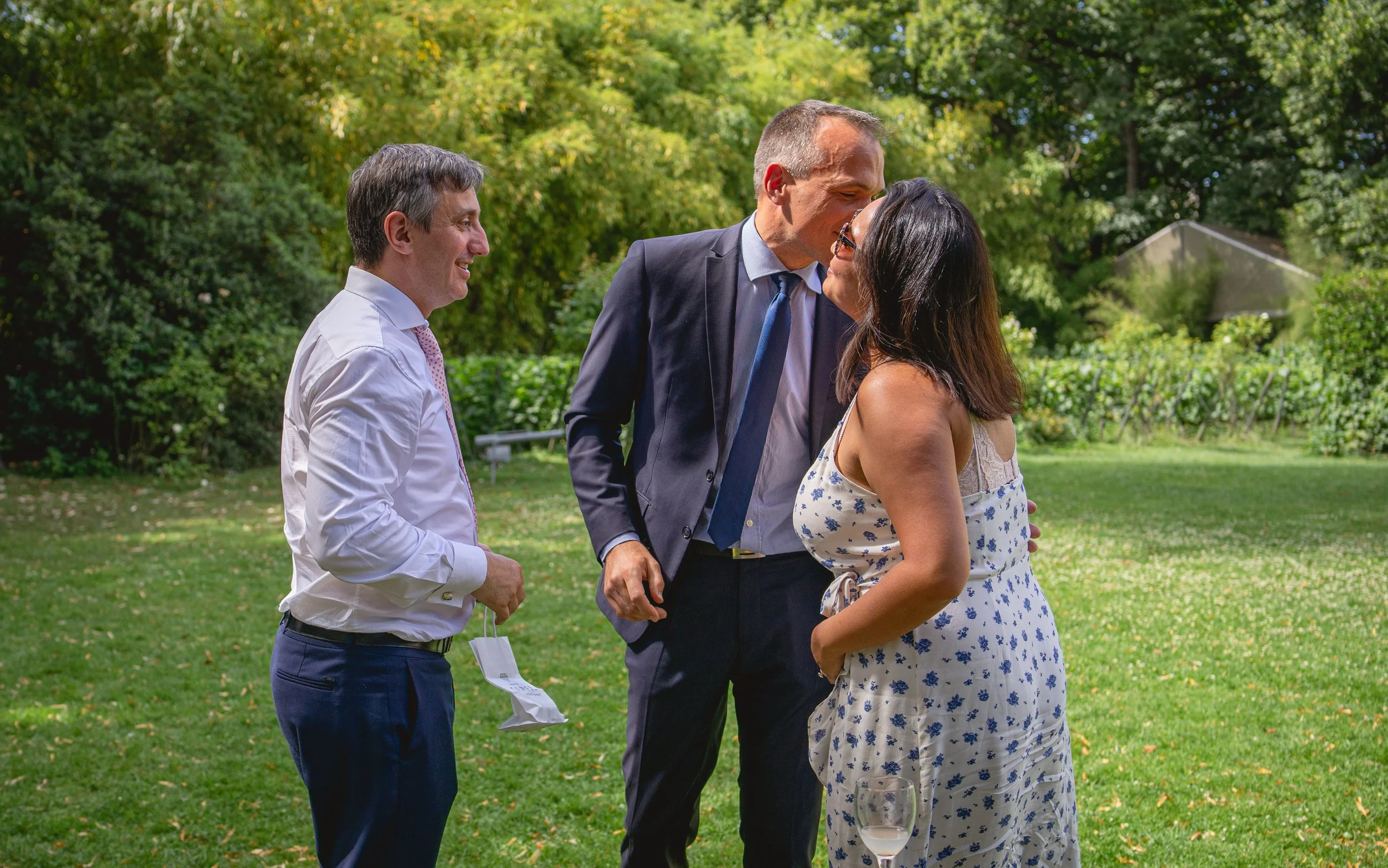 Un groupe de trois personnes, deux hommes et une femme, en train de discuter dans un jardin ensoleillé. L'homme au centre et la femme semblent s'embrasser ou sont très proches, tandis que l'autre homme sourit en regardant.