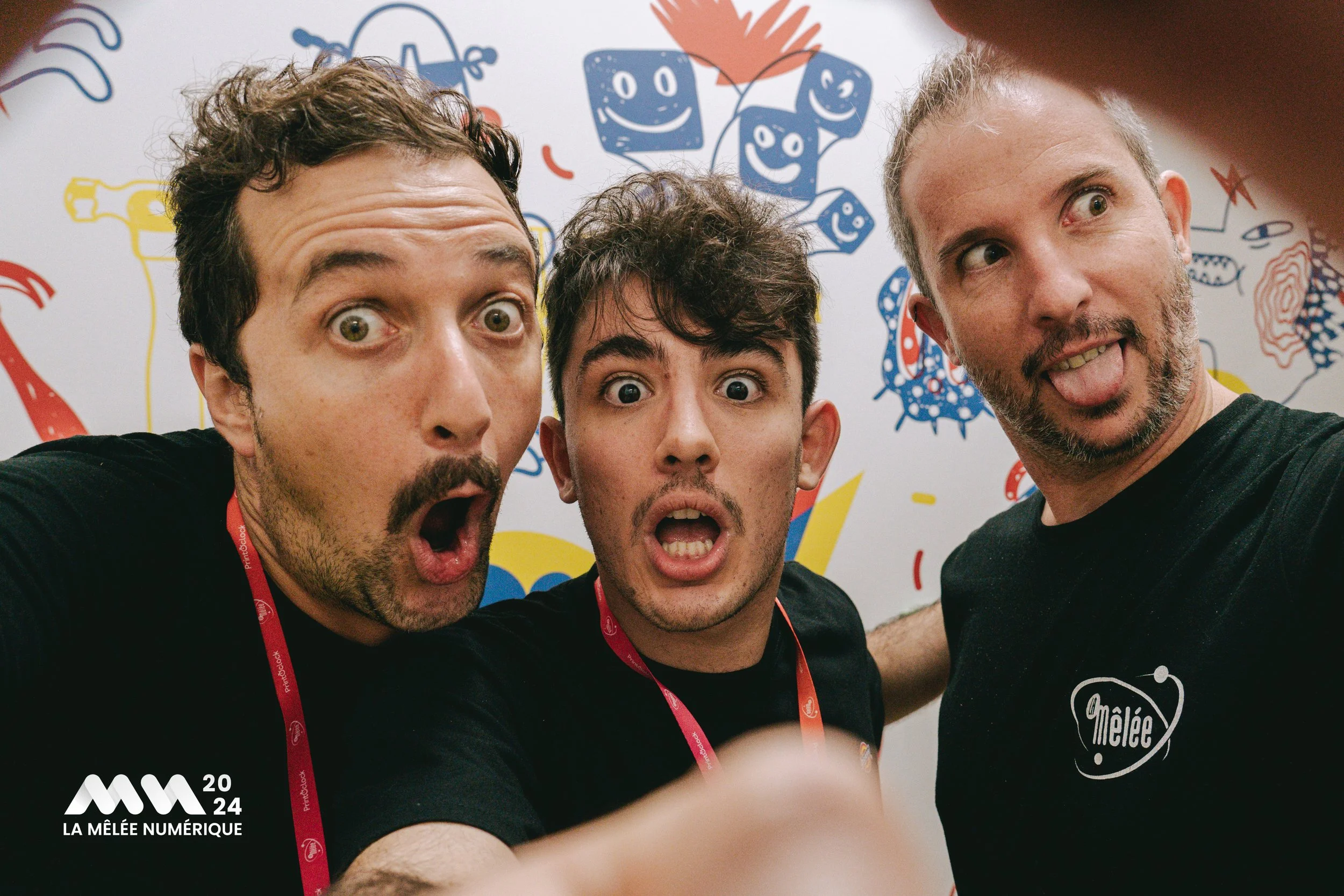 Trois hommes prennent un selfie devant un mur décoré avec des dessins colorés de personnages et d'objets. Ils portent des t-shirts noirs avec un logo 'Mêlée'.