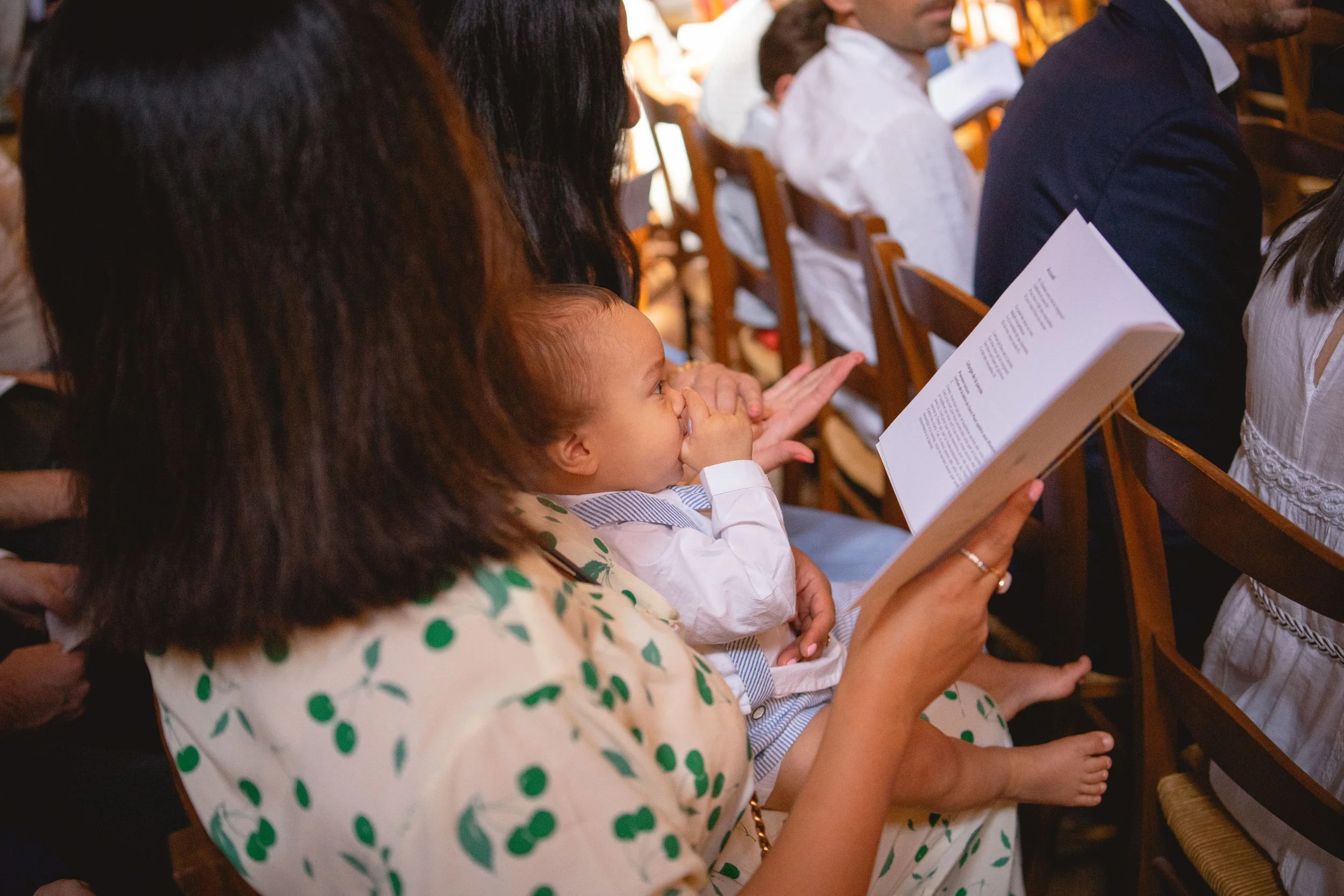 Un jeune enfant assis sur les genoux d'une femme dans une salle, regardant un programme ou un livre, avec d'autres personnes assises à côté lors d'un événement ou une réunion.
