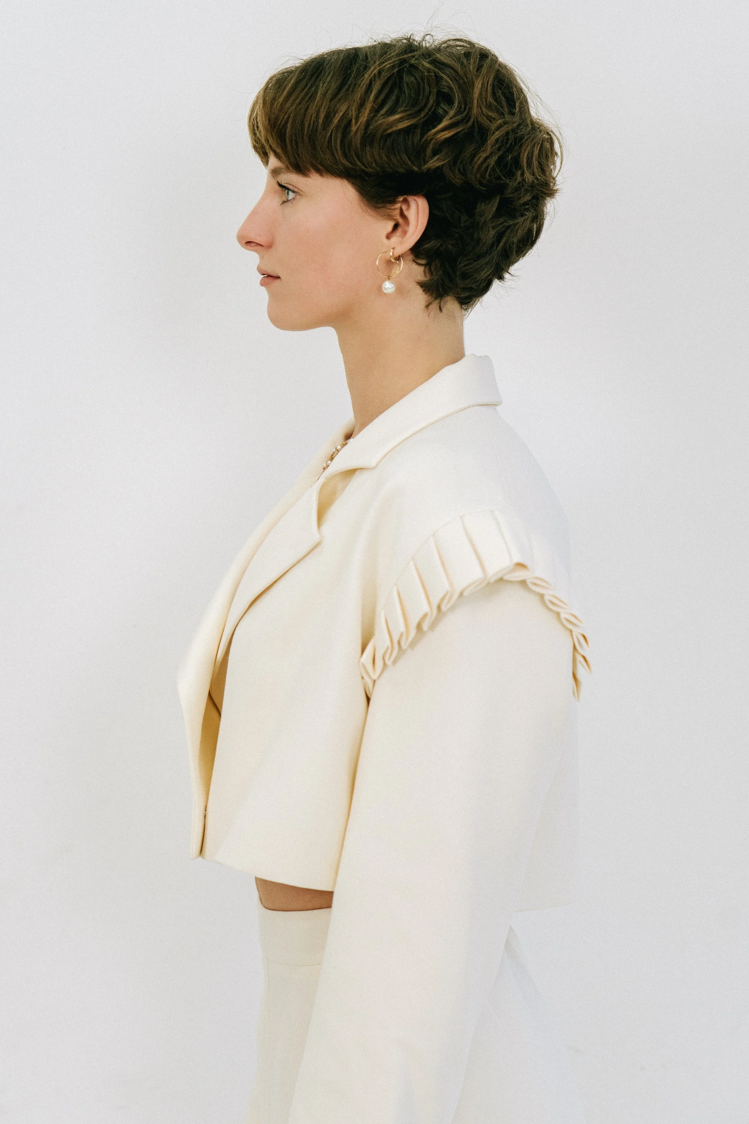 Profil d'une femme aux cheveux courts bruns, portant une veste blanche et des boucles d'oreilles perlées, contre un fond blanc.