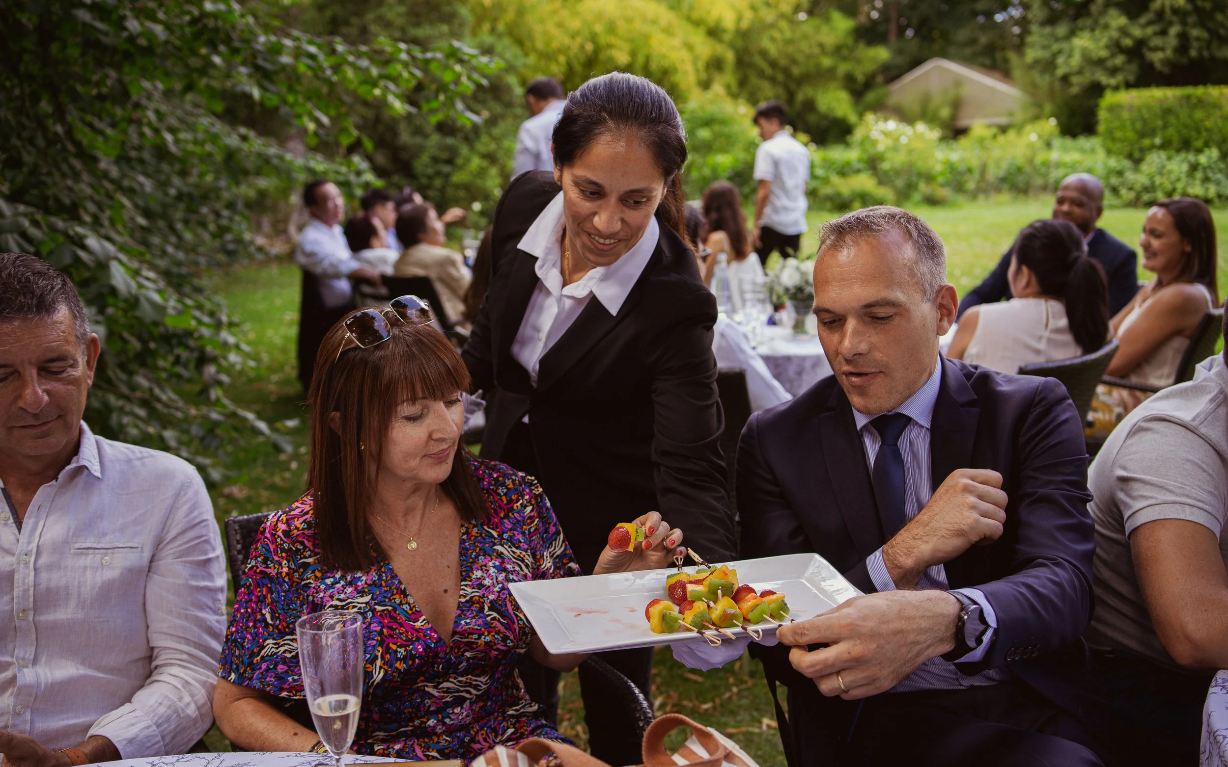 Groupe de personnes lors d'un repas en plein air, avec un serveur servant des amuse-bouches à un homme en costume.
