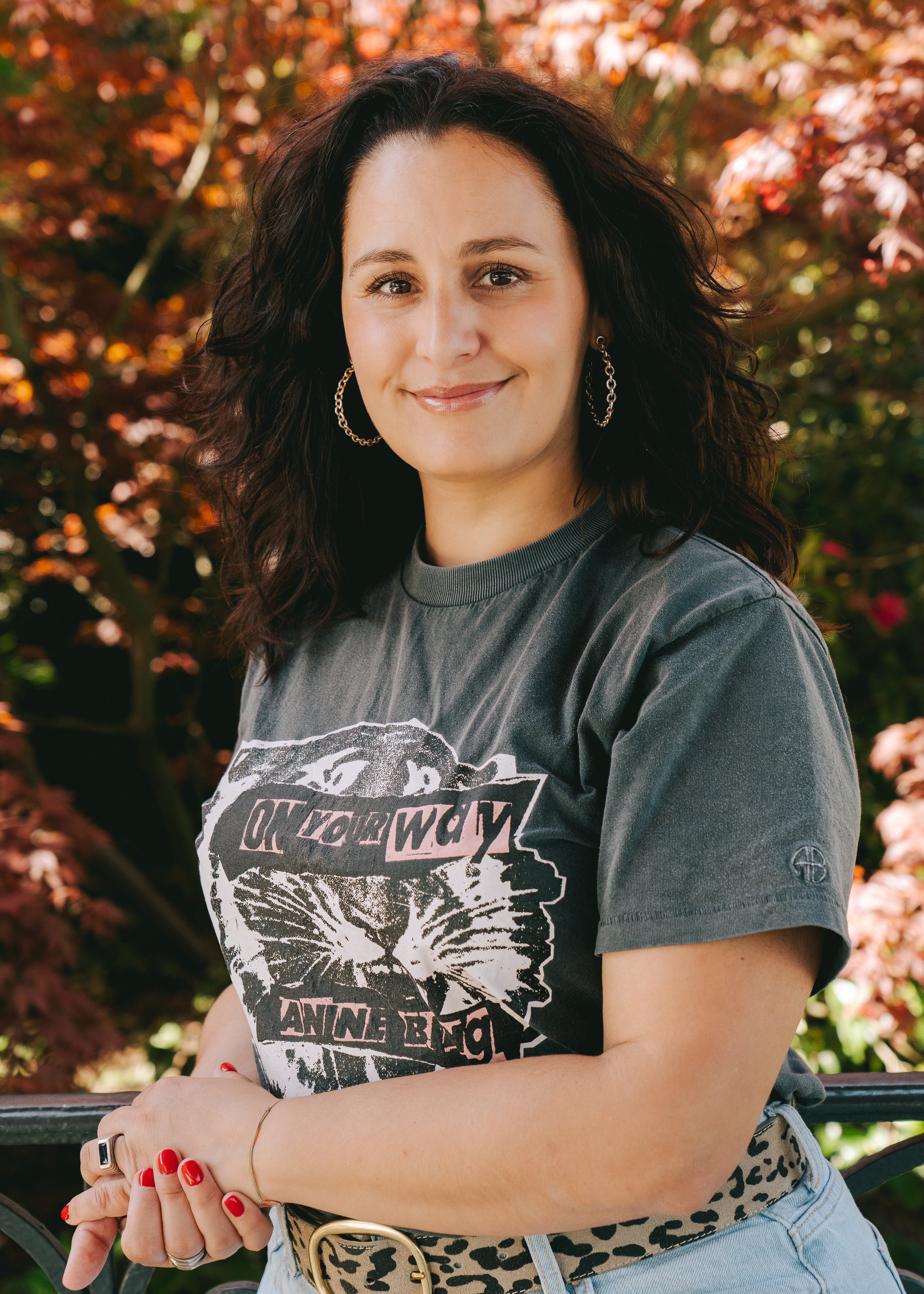 Une femme souriante avec des cheveux bouclés, portant un t-shirt gris avec un motif de chat, des boucles d'oreilles en cercle, et une ceinture à motif léopard, posant devant un décor d'automne avec des feuilles orange et rouges.