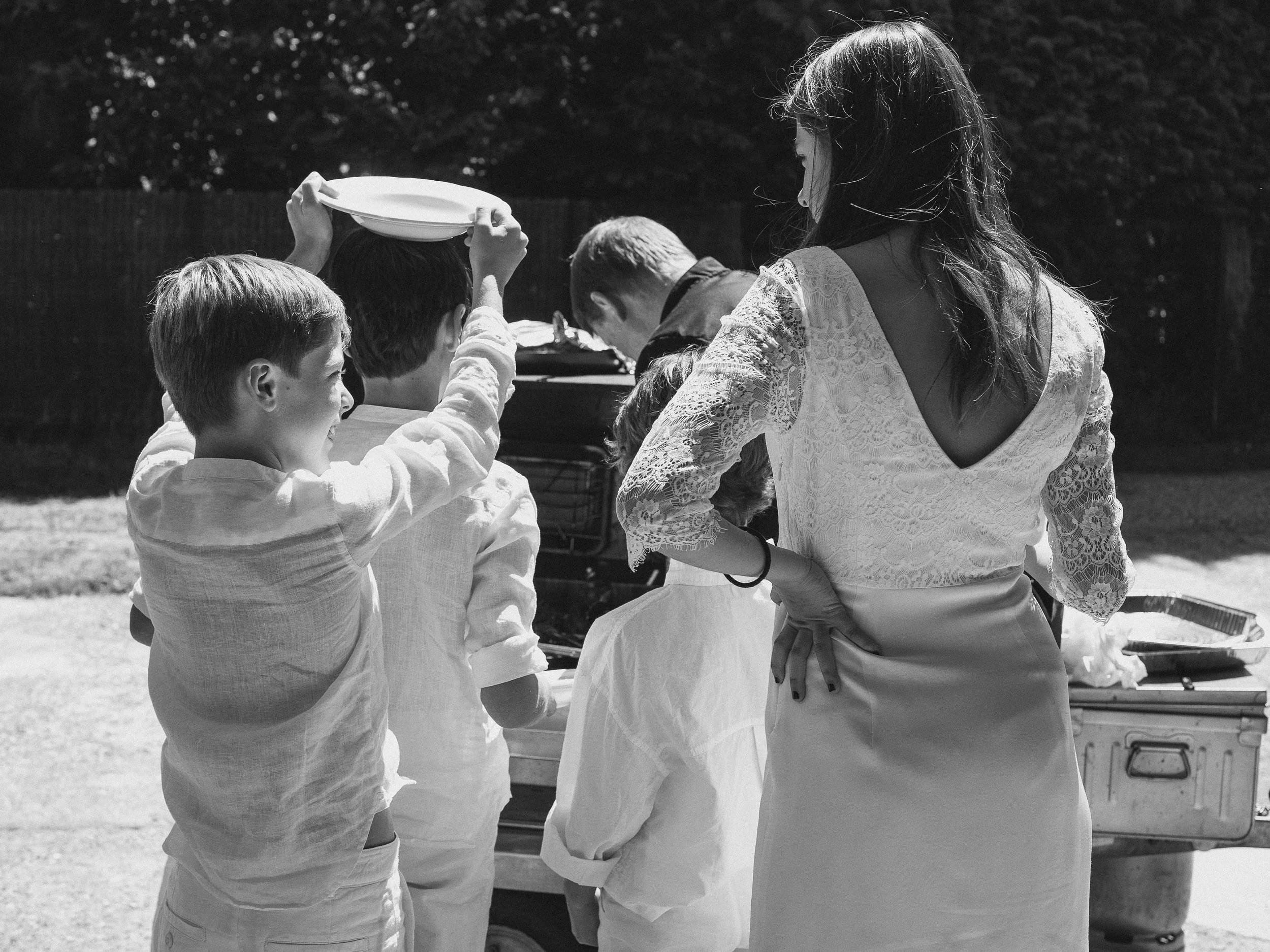 Groupe de personnes, principalement des enfants, de dos, en extérieur lors d’un repas ou pique-nique. Une femme en robe blanche avec un dos nu regarde vers le groupe, un garçon avec une chemise claire tient un plat au-dessus de sa tête, d’autres enfa