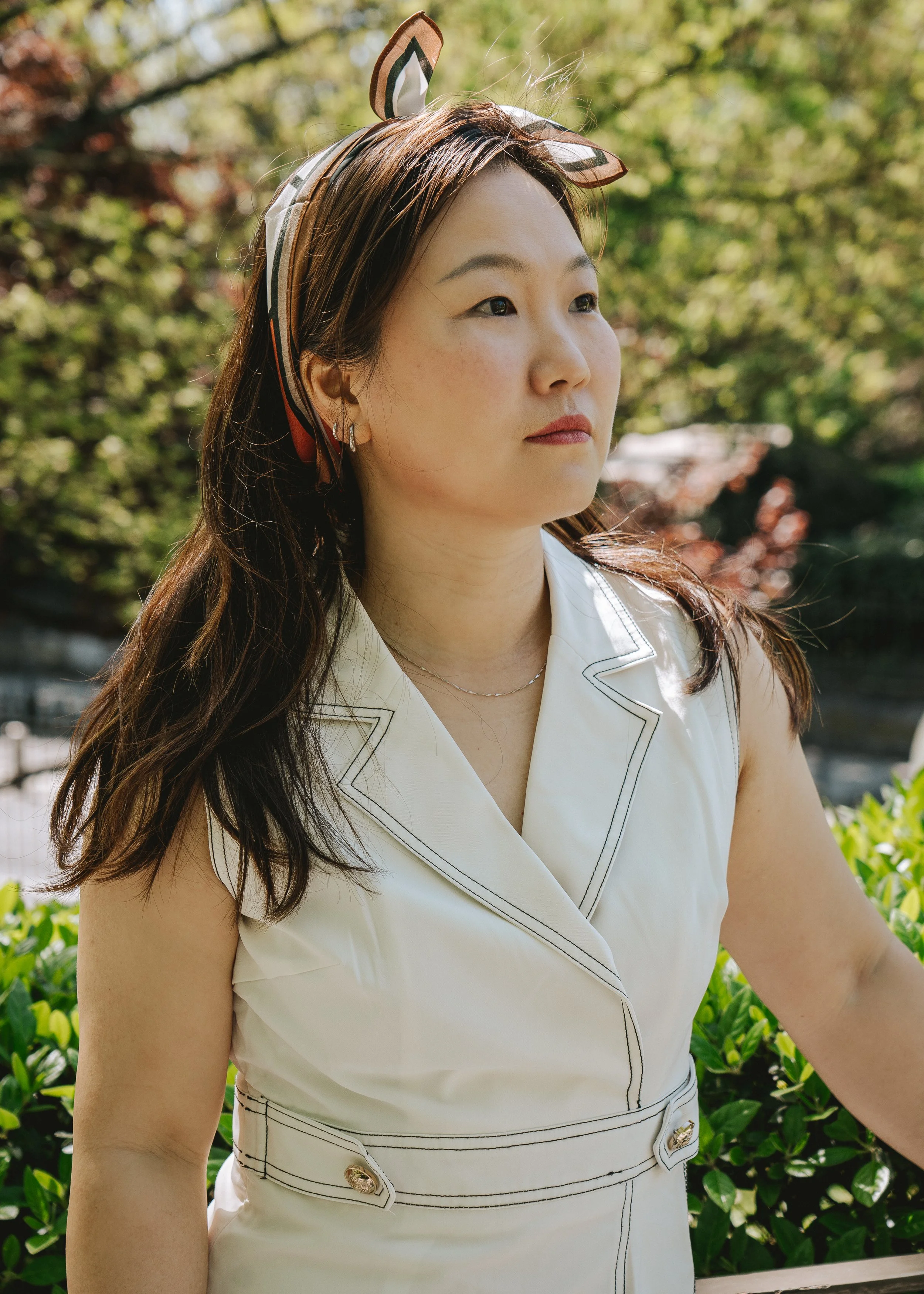 Une femme asiatique porte une tenue blanche avec des détails noirs, une bande à cheveux avec des oreilles de lapin, et se tient devant des arbres en plein air.