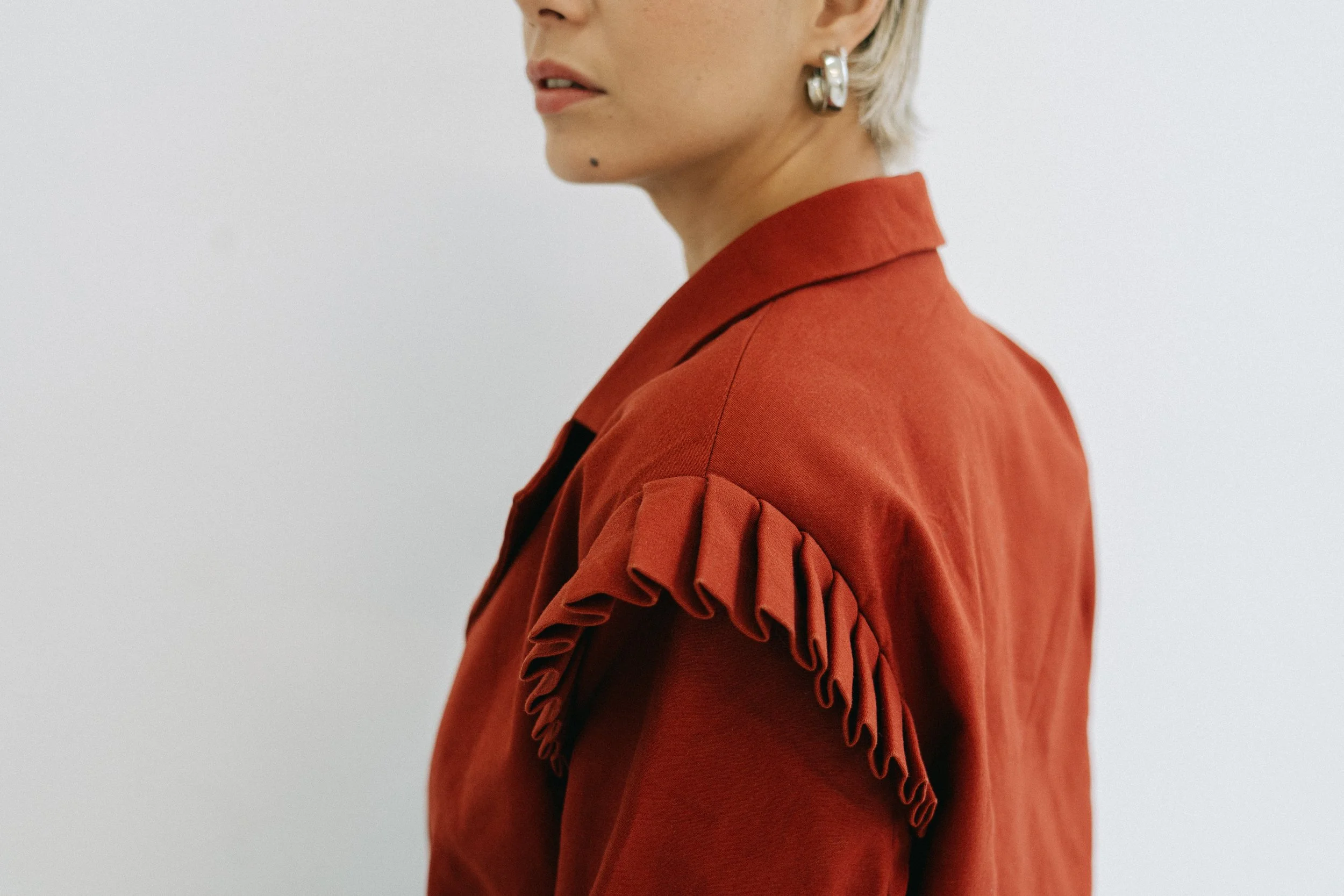 Une femme avec des cheveux blonds courts, portant une veste rouge avec des détails en volants sur les épaules, regarde de côté contre un fond blanc.