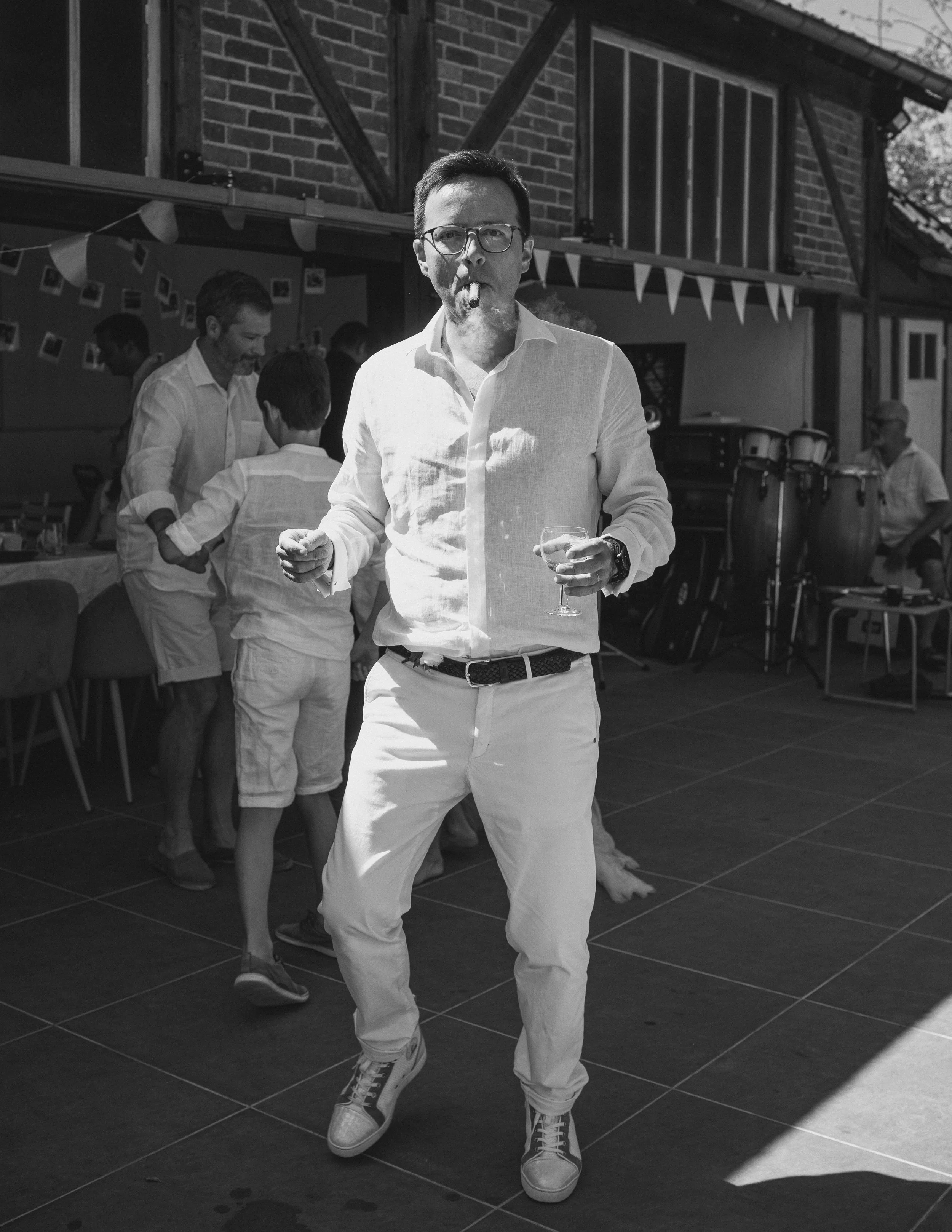 Un homme portant des lunettes, en chemise blanche et pantalon clair, danse avec une boisson à la main lors d'une fête en extérieur, avec des autres invités en arrière-plan.
