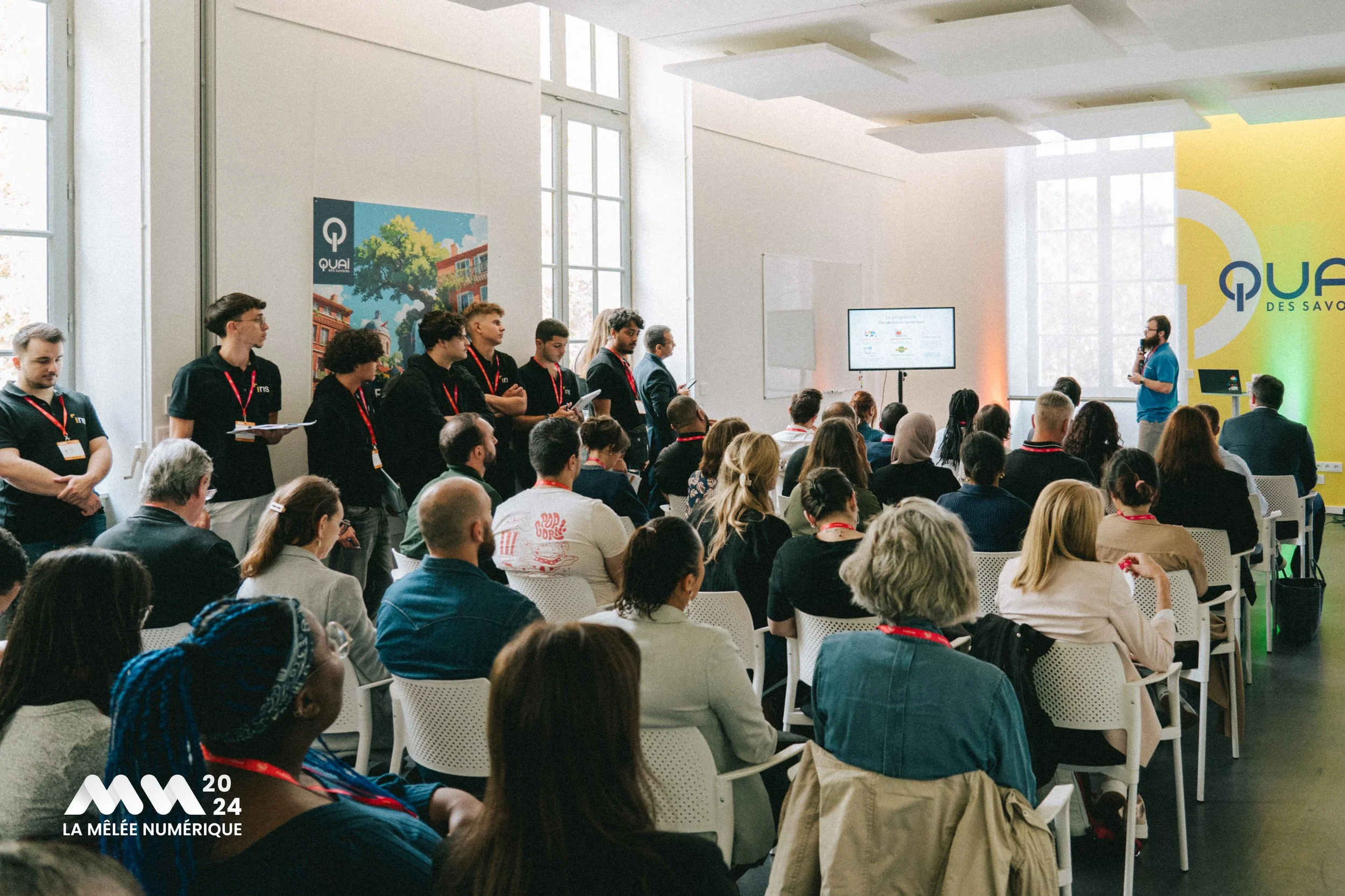 Une conférence avec un public et un groupe de personnes en noir à l'arrière gauche. Un conférencier parle à une audience dans une salle lumineuse avec des grandes fenêtres. Logo 'Qual' et 'La Mêlée Numérique 2024' dans le coin inférieur gauche.