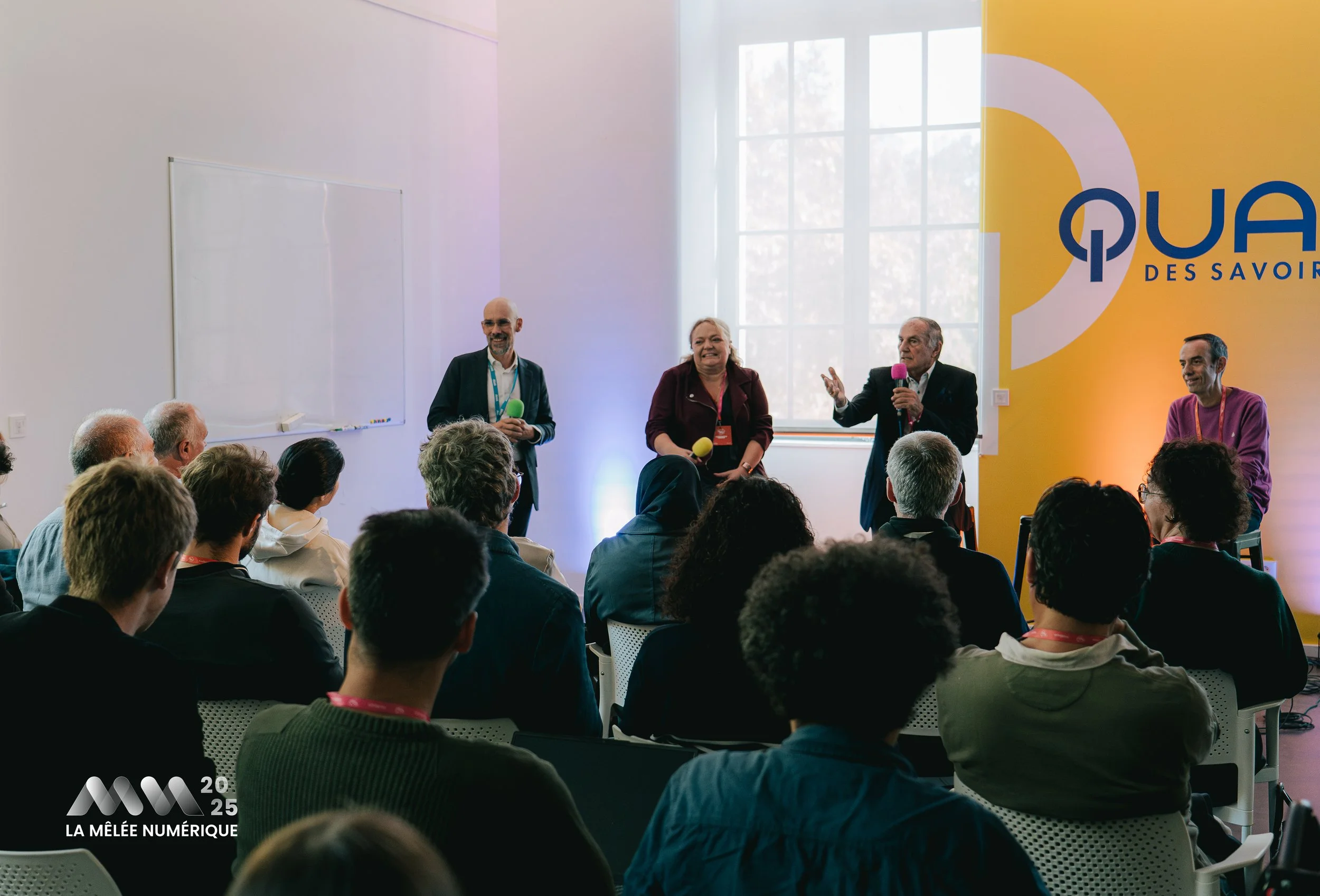 Une conférence avec cinq intervenants debout devant un public assis, dans une salle lumineuse avec une grande fenêtre. Un panneau jaune à l'arrière affiche un logo et le texte « QUAI DES SAVOIR ».