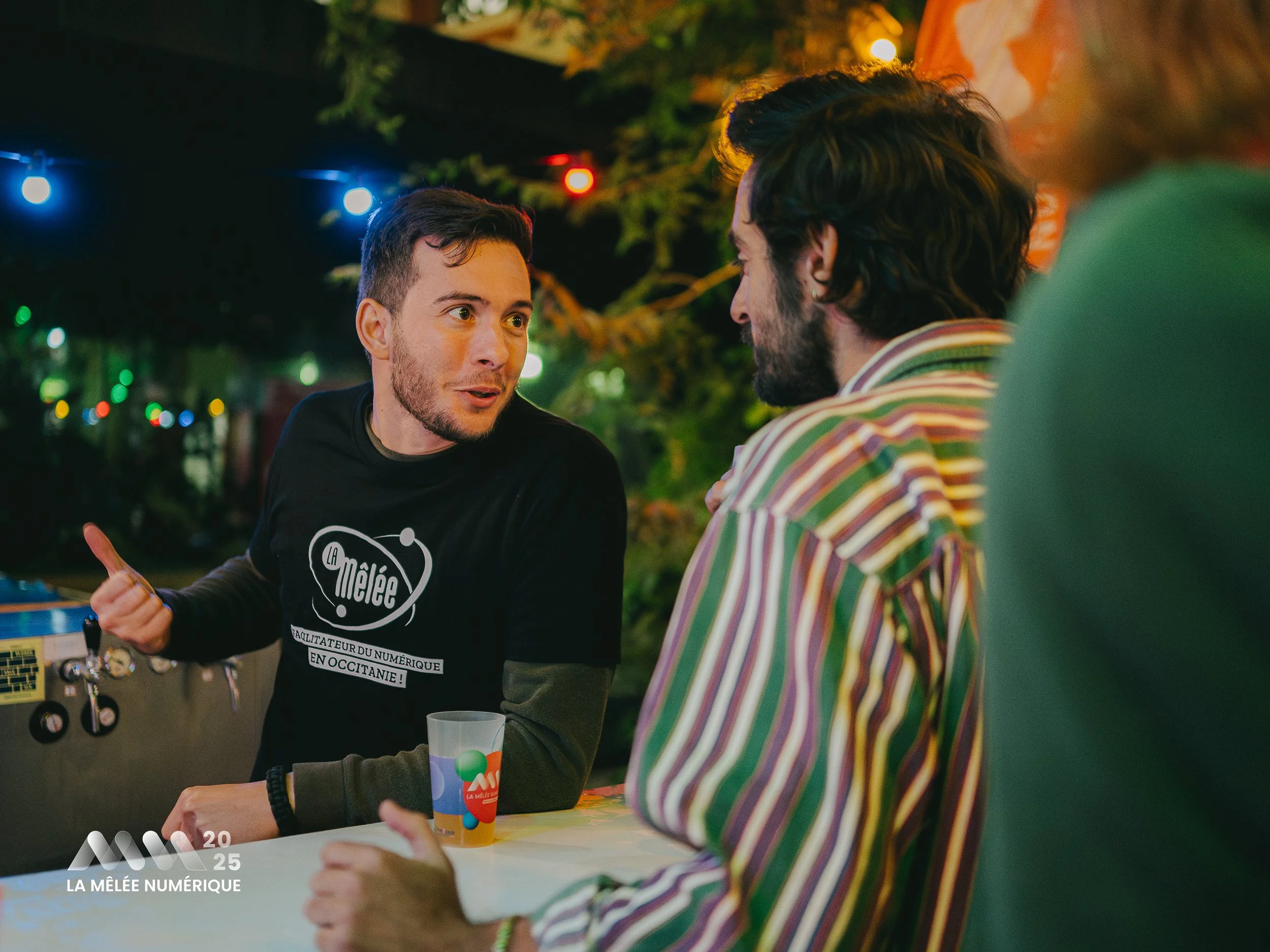 Deux hommes discutent à une table lors d'une soirée en extérieur, avec des lumières colorées en arrière-plan. L'un porte un T-shirt noir avec un logo et un texte, l'autre est habillé d'une chemise rayée.