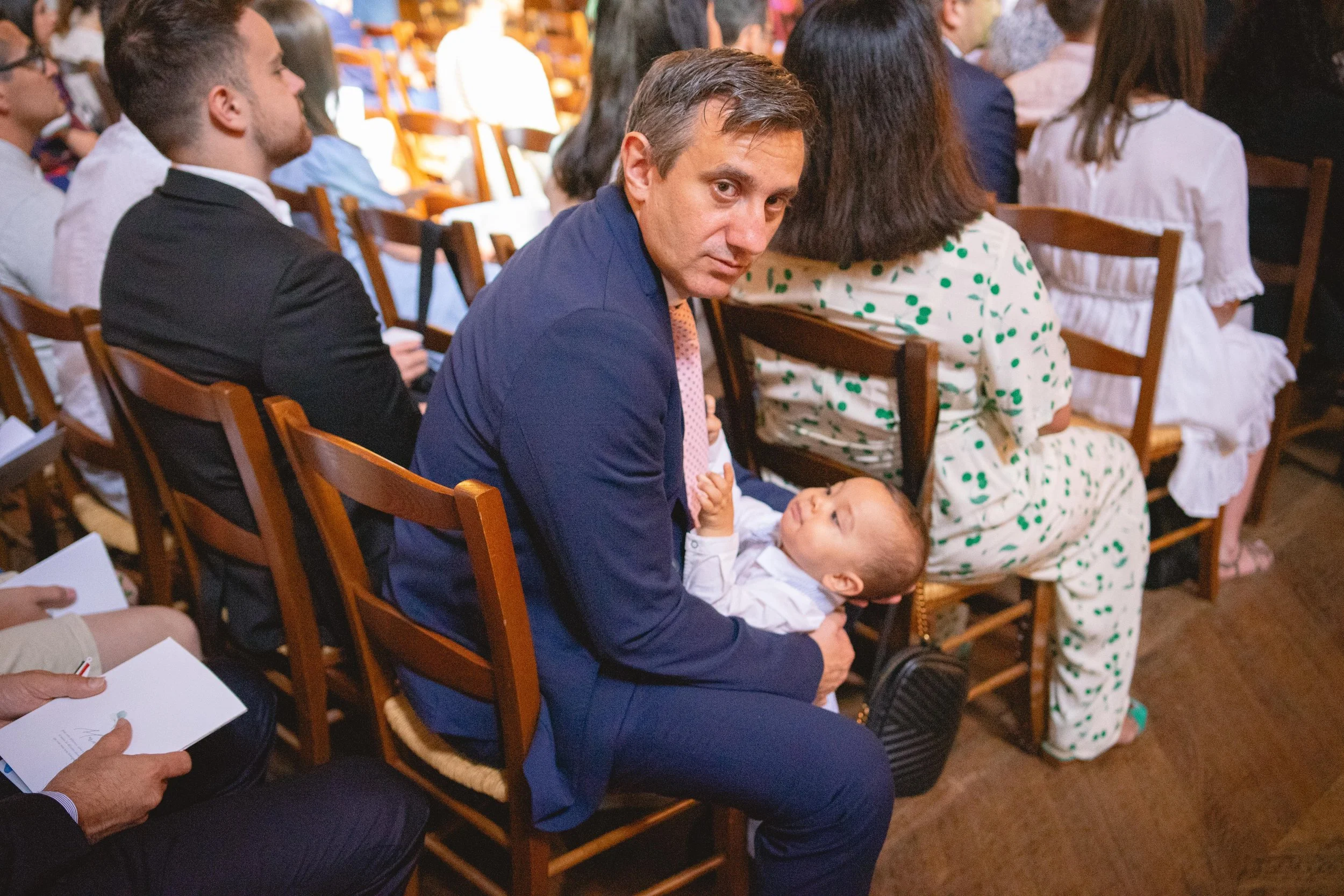 Un homme en costume bleu assis dans une salle pleine de personnes, tenant un bébé dans ses bras, regardant vers l'appareil photo.