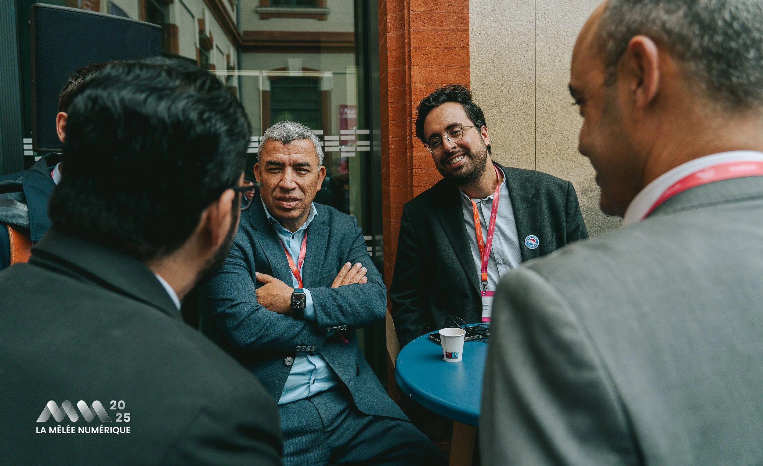 Groupe de quatre hommes en costume discutant autour d'une table lors de La Mêlée Numérique 2025, l'un d'eux porte une montre connectée.
