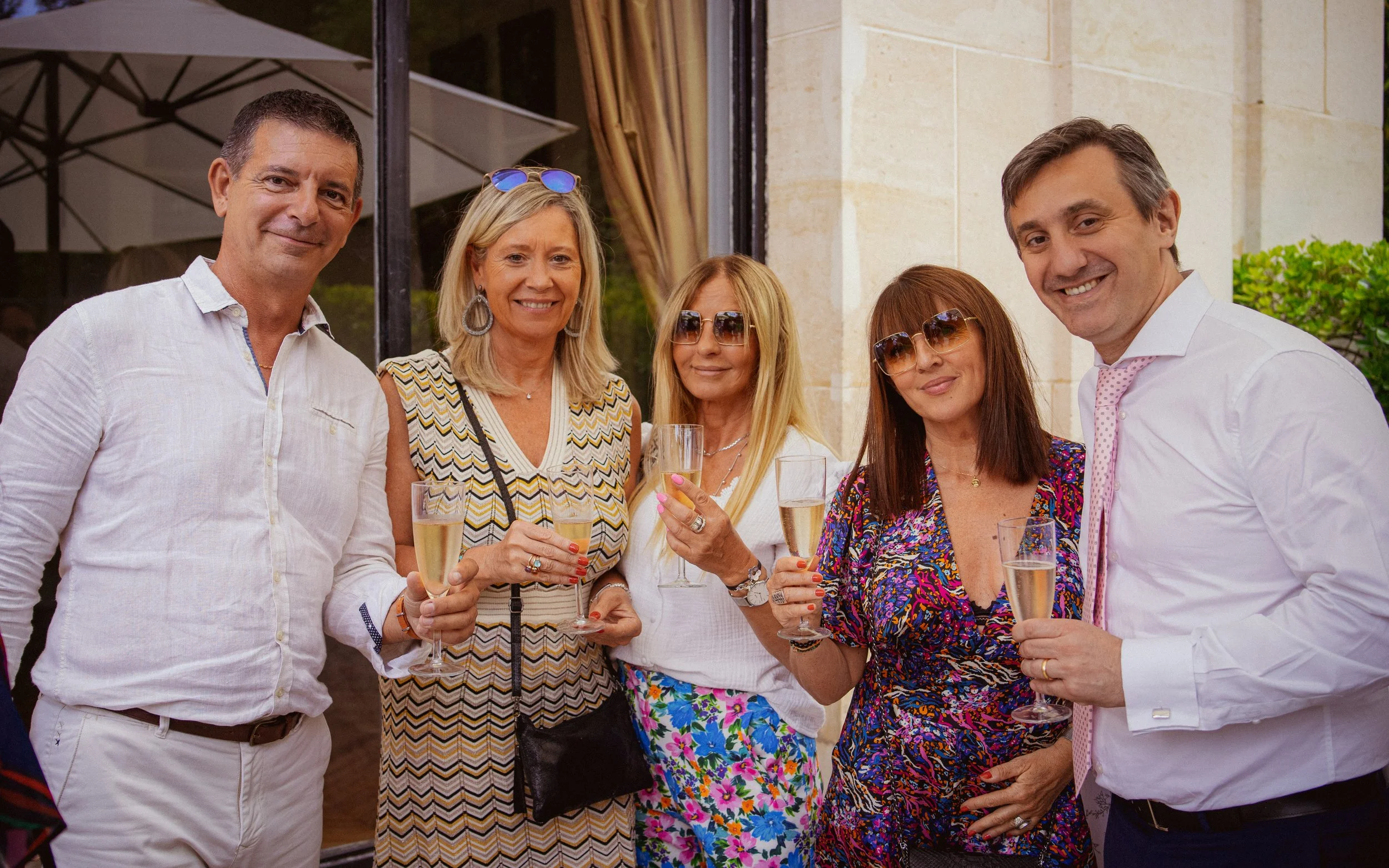 Cinq personnes souriantes tenant des verres de champagne lors d'une fête en extérieur.