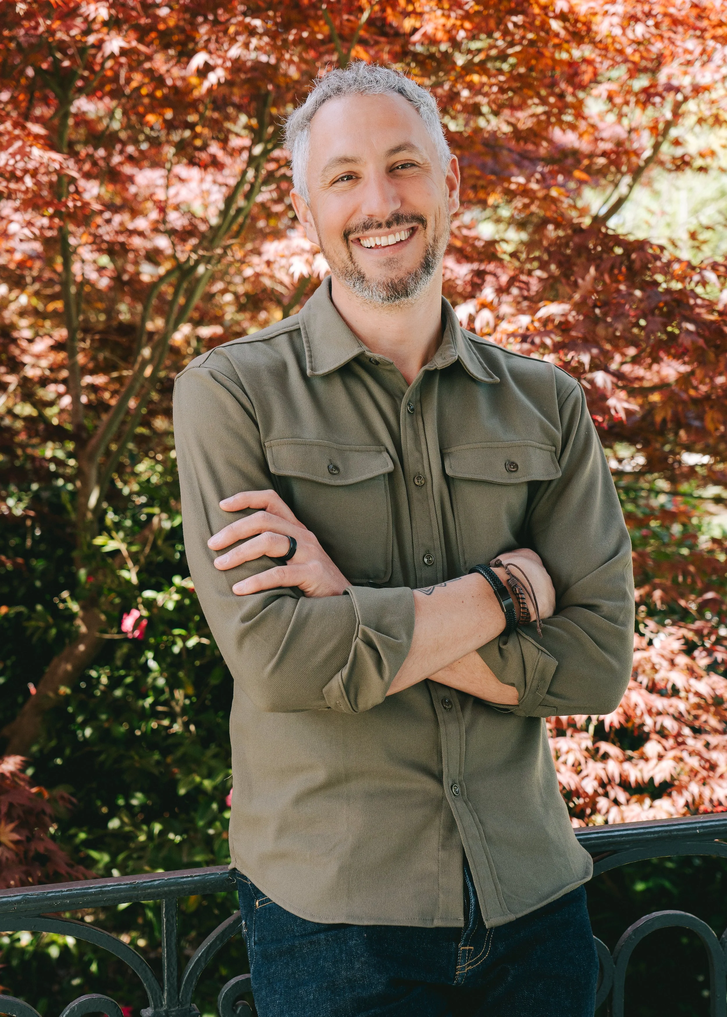 Un homme souriant avec des cheveux gris et une barbe, portant une chemise kaki, se tenant avec les bras croisés devant un arbre aux feuilles rouges en automne.