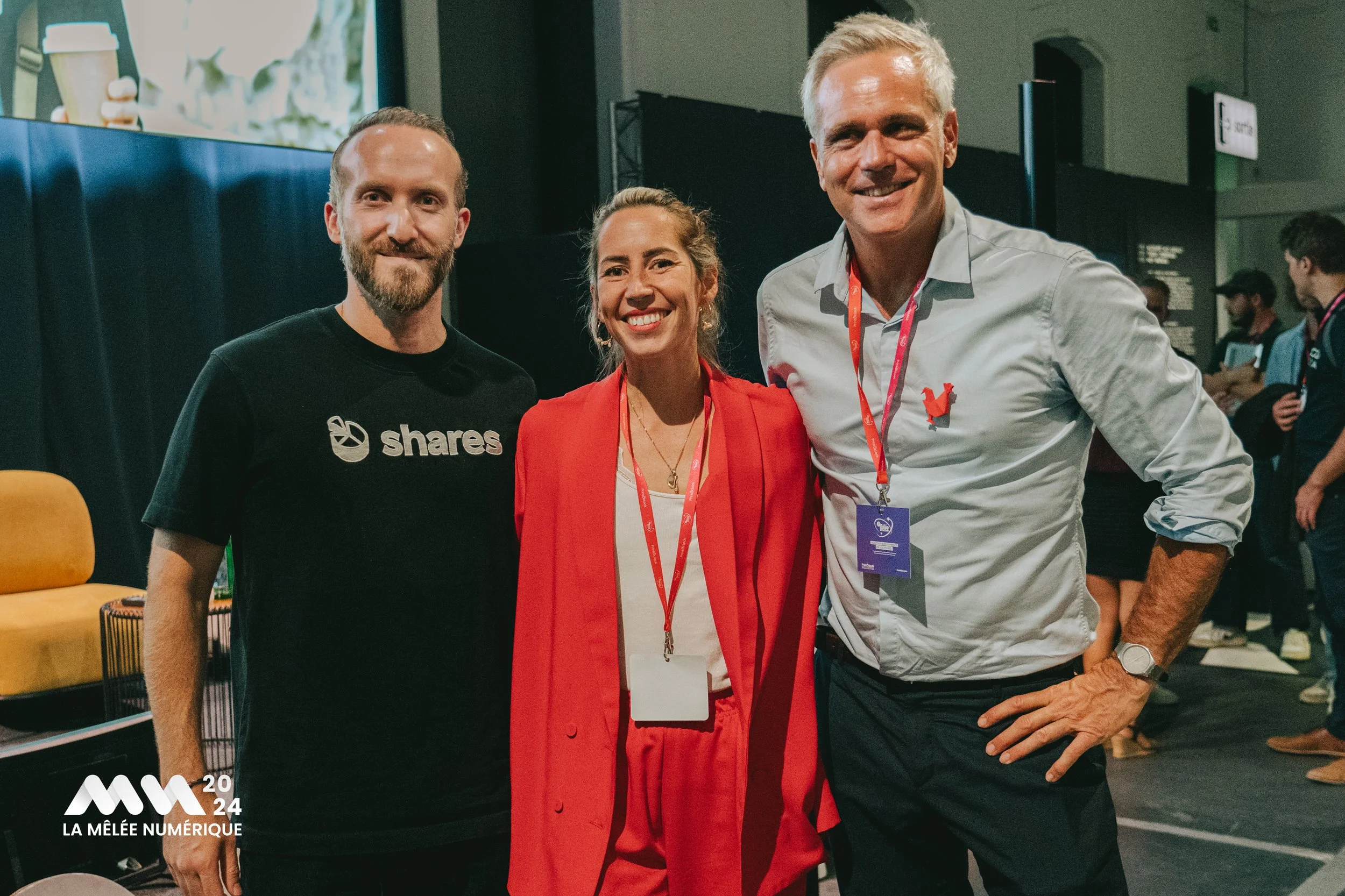 Trois personnes souriantes posent ensemble lors d'une conférence ou événement numérique, avec un fond sombre et un écran visible. Une femme en costume rouge au centre, un homme en t-shirt noir à gauche et un homme en chemise blanche à droite.