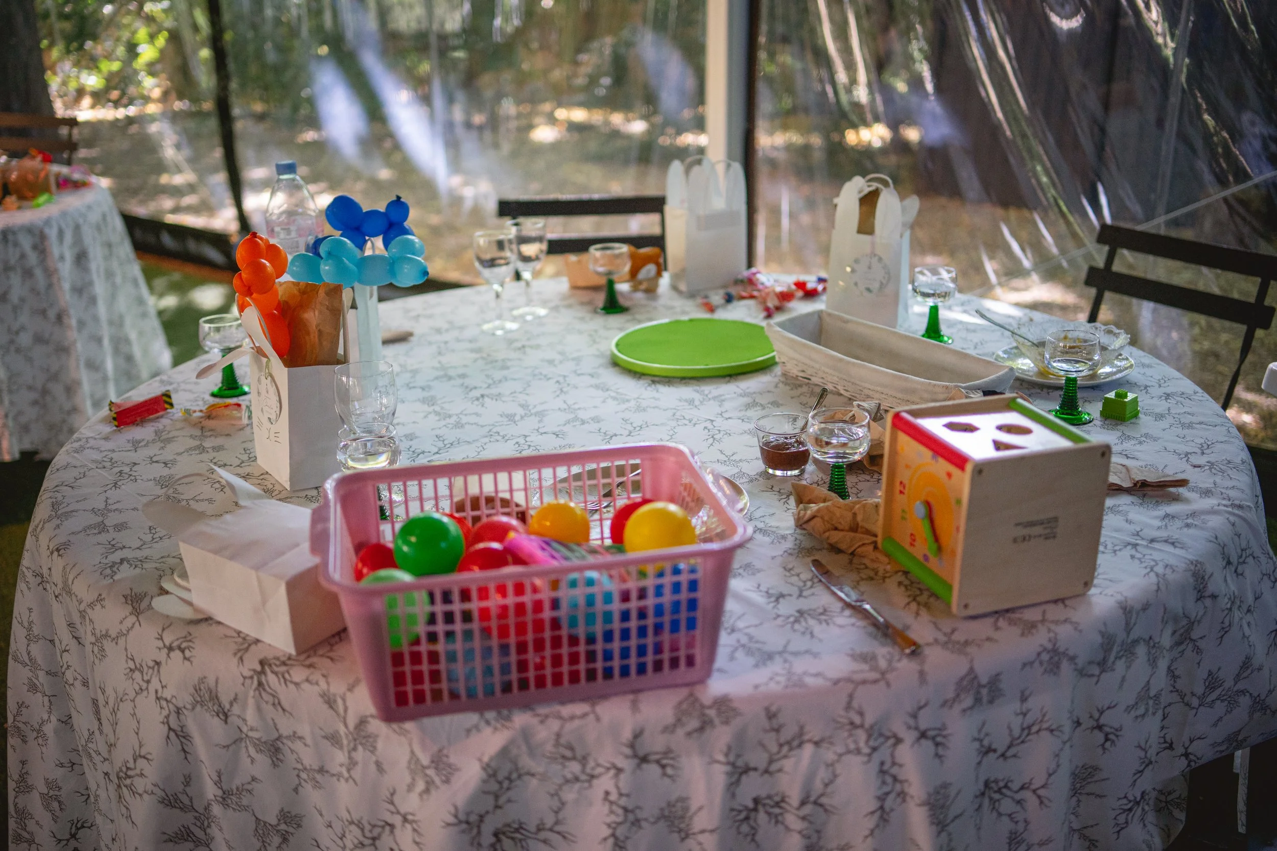Table de fête décorée avec des ballons, jouets, verres d'eau, et des paniers de cadeaux dans un environnement lumineux.