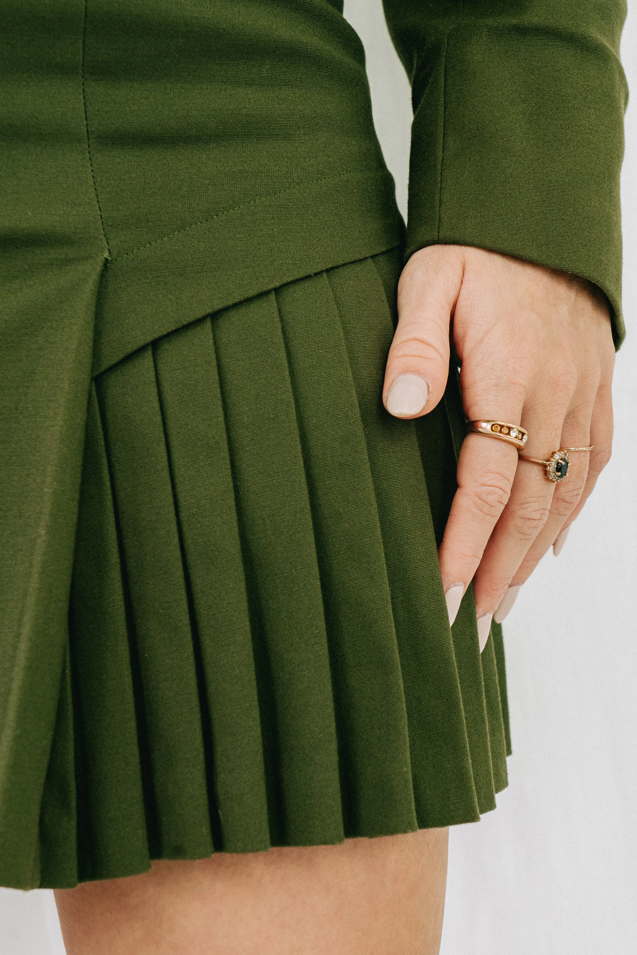 Une personne porte une jupe plissée vert foncé et un manteau assorti, avec des mains ornées de bagues.