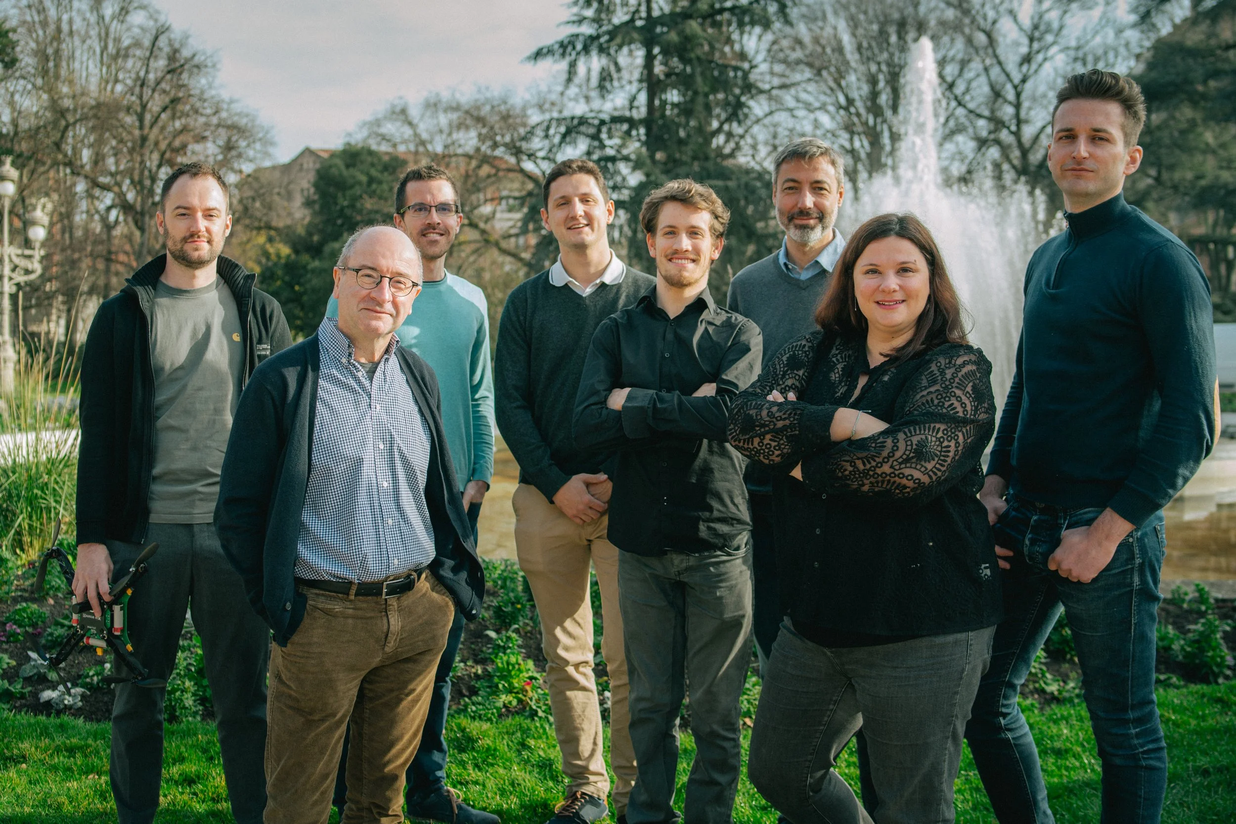 Groupe de neuf personnes posant en extérieur près d'une fontaine dans un parc, avec des arbres en arrière-plan, lors d'une journée ensoleillée.