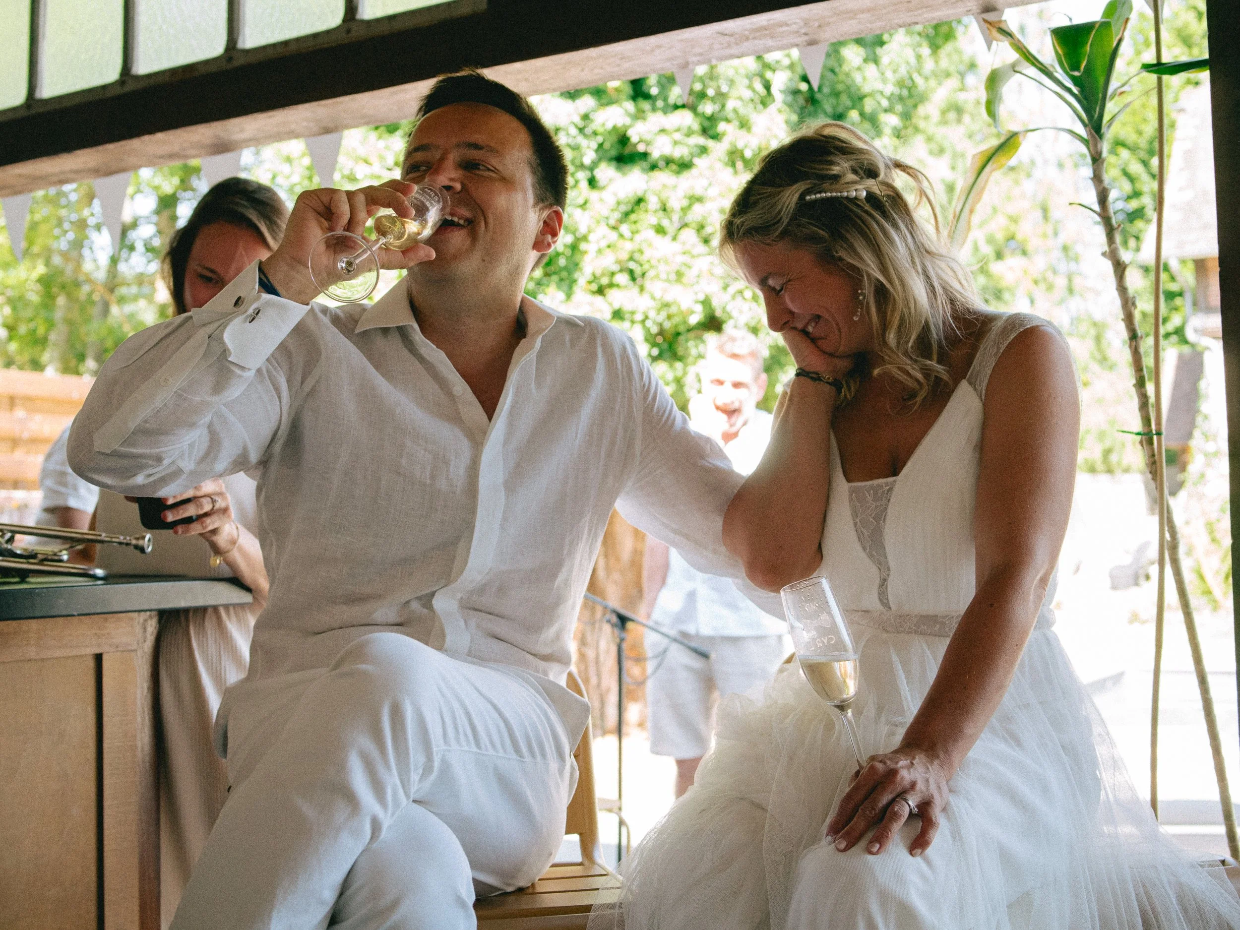 Un groupe de personnes lors d'une célébration en plein air. Une femme en robe blanche en pleurs, appuyée sur une table, et un homme en chemise blanche souriant et buvant du champagne, avec d'autres personnes en arrière-plan, dans un environnement ver