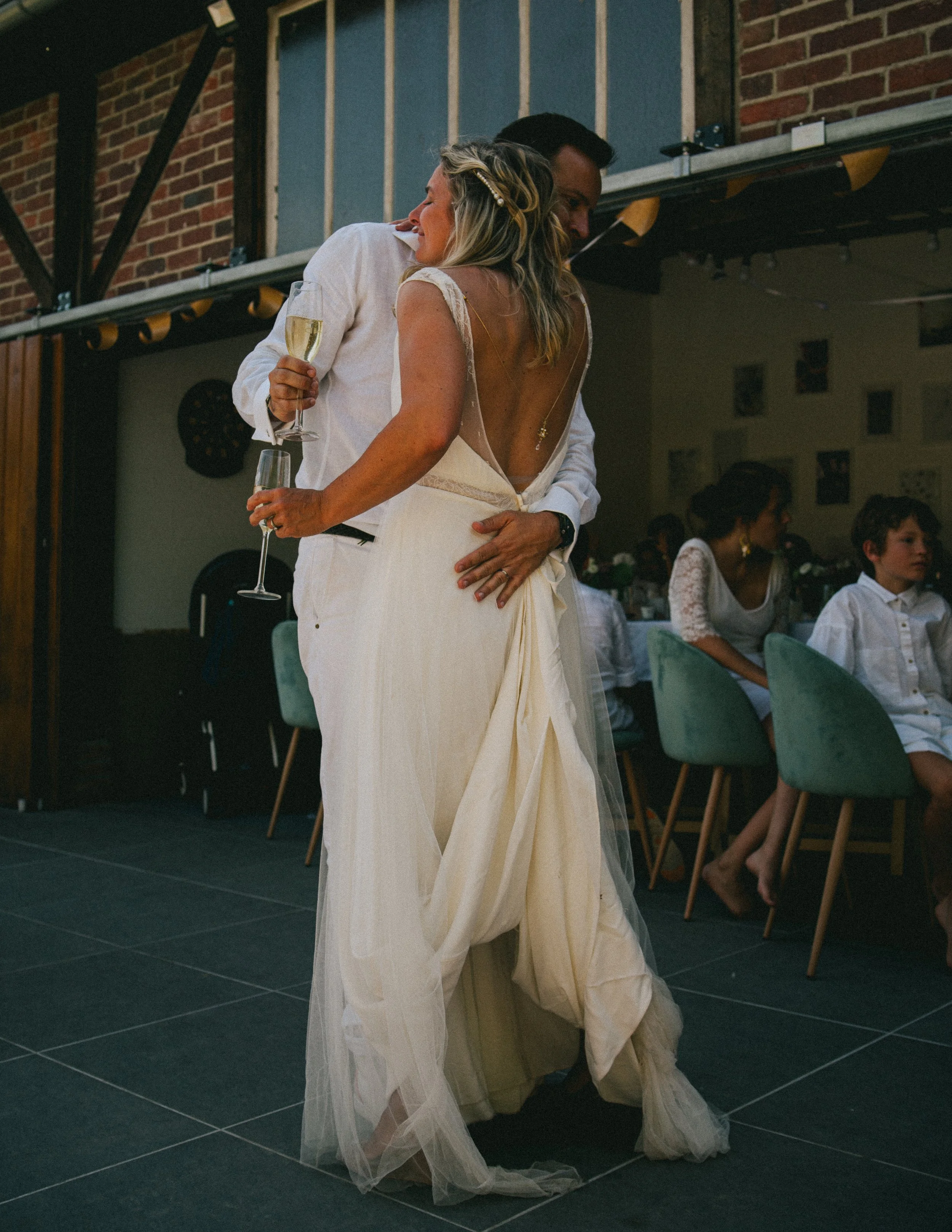 Un couple danse lors d'un mariage, la femme en robe de mariée blanche et le homme en chemise blanche, avec une coupe de champagne à la main, dans une salle décorée pour l'événement.