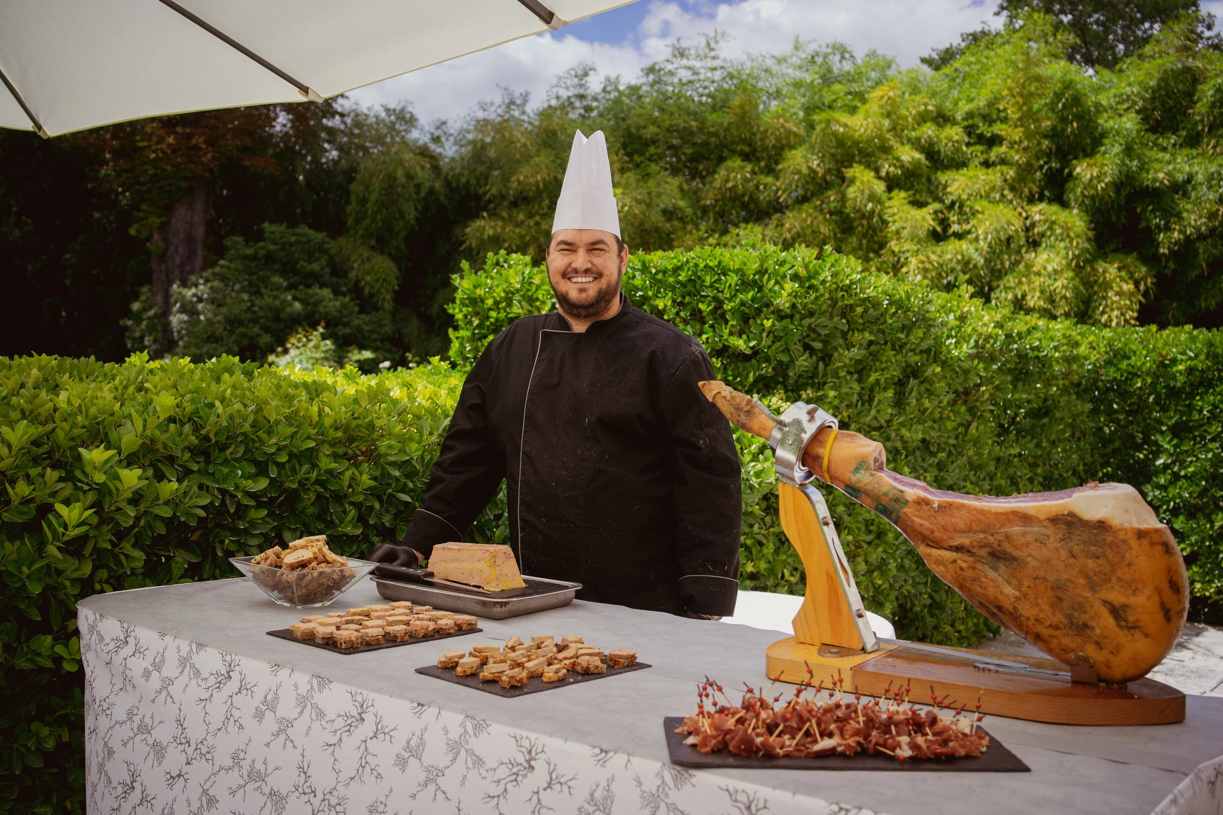 Homme souriant en tenue de chef, debout derrière une table, entouré de nourriture, avec un jambon cru suspendu à une machine à jambon, en extérieur avec des arbres en arrière-plan.