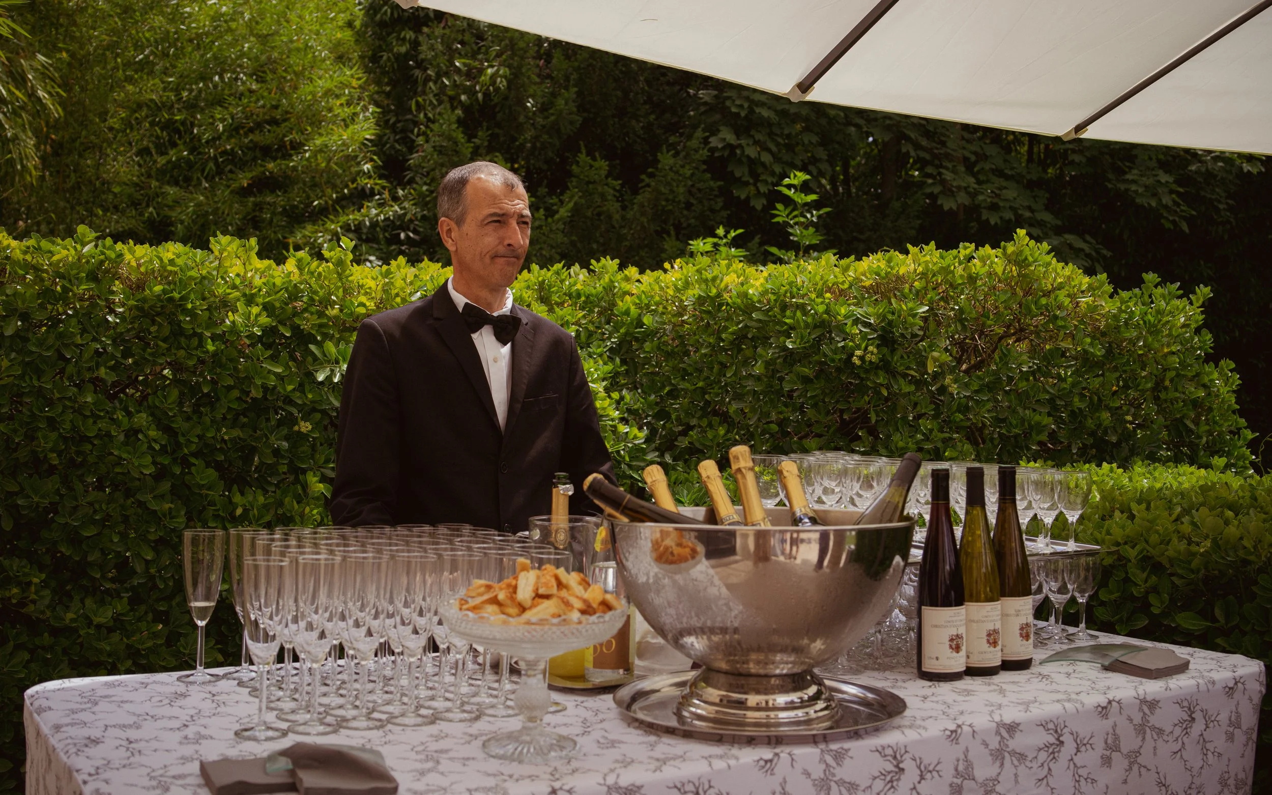Hommes en costume noir servant un buffet de champagne et amuse-gueules sous un parasol dans un jardin.