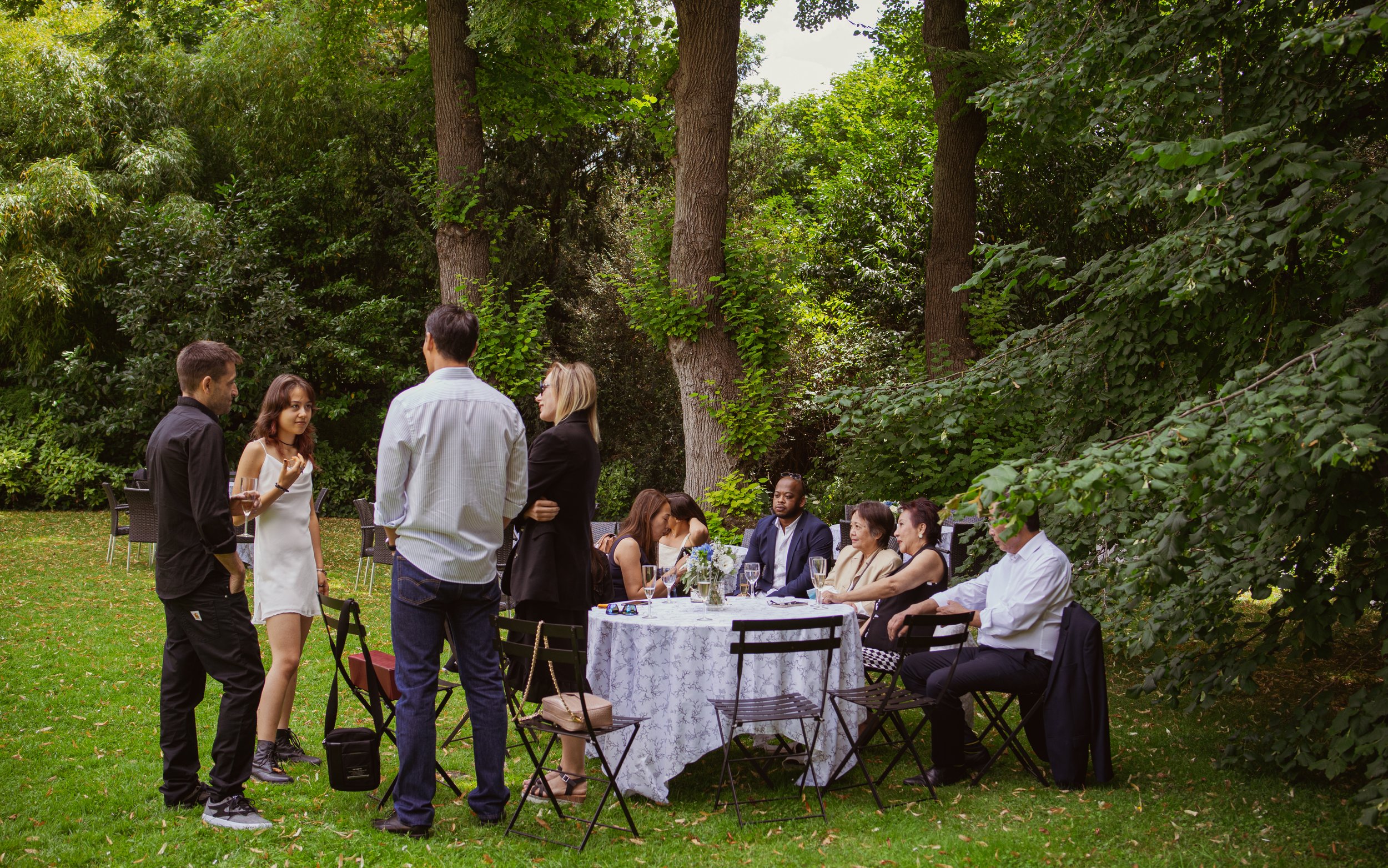 Groupe de personnes à un dîner en plein air dans un jardin verdoyant, certains discutant debout, d'autres assis autour d'une table avec une nappe blanche et des fleurs.