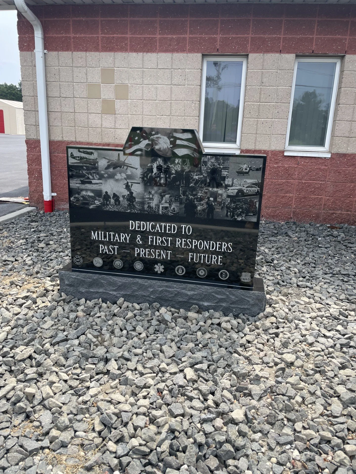 Black commemorative plaque dedicated to military and first responders, with images of helicopters, soldiers, police, firefighters, and emergency personnel, on a bed of gray rocks outside a building.