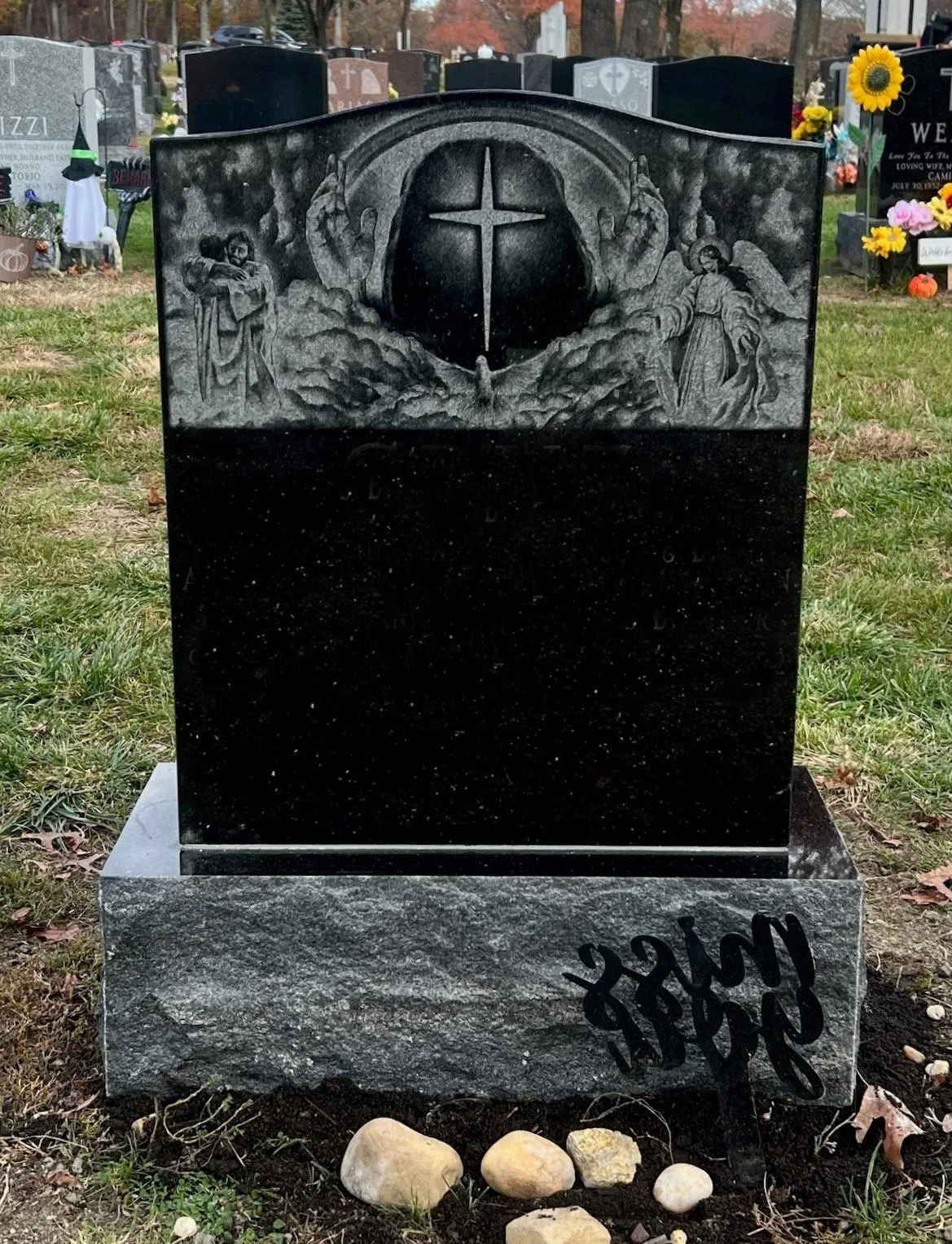 A black granite headstone with engraved religious imagery, including angels, clouds, and a cross. The lower part of the headstone is blank, and there is an inscription on the base reading 'In Loving Memory' with a few rocks placed in front.