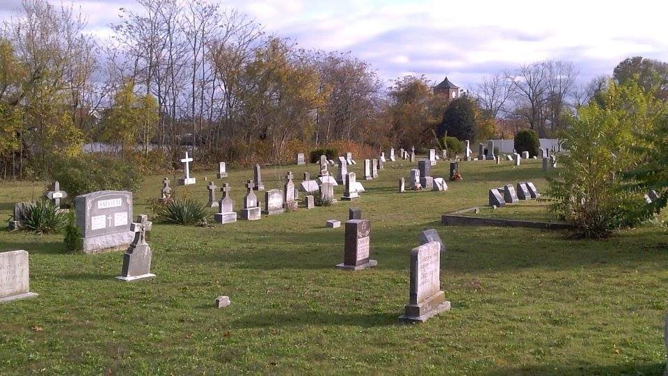 A cemetery with numerous gravestones and crosses, surrounded by trees with autumn foliage and a cloudy sky.