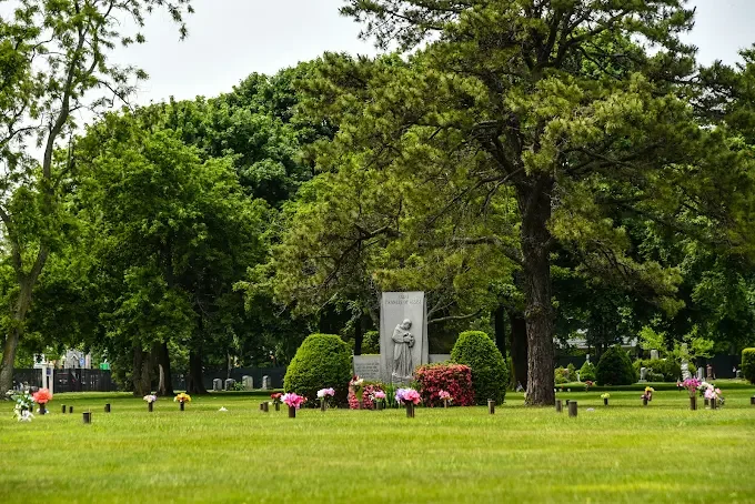 A peaceful cemetery scene with lush green grass, several trees, and a large stone monument surrounded by colorful flowers and potted plants.